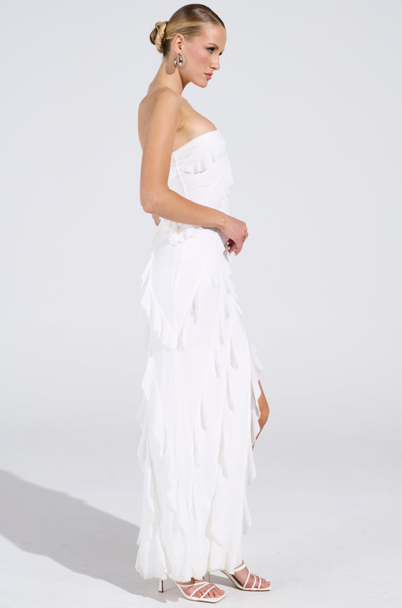 Side view of a person in an ELLA MESH TUBE DRESS IN WHITE with layered fringes. They have a neat bun, hoop earrings, and high-heeled sandals. The simple light backdrop emphasizes the dress's elegance.
