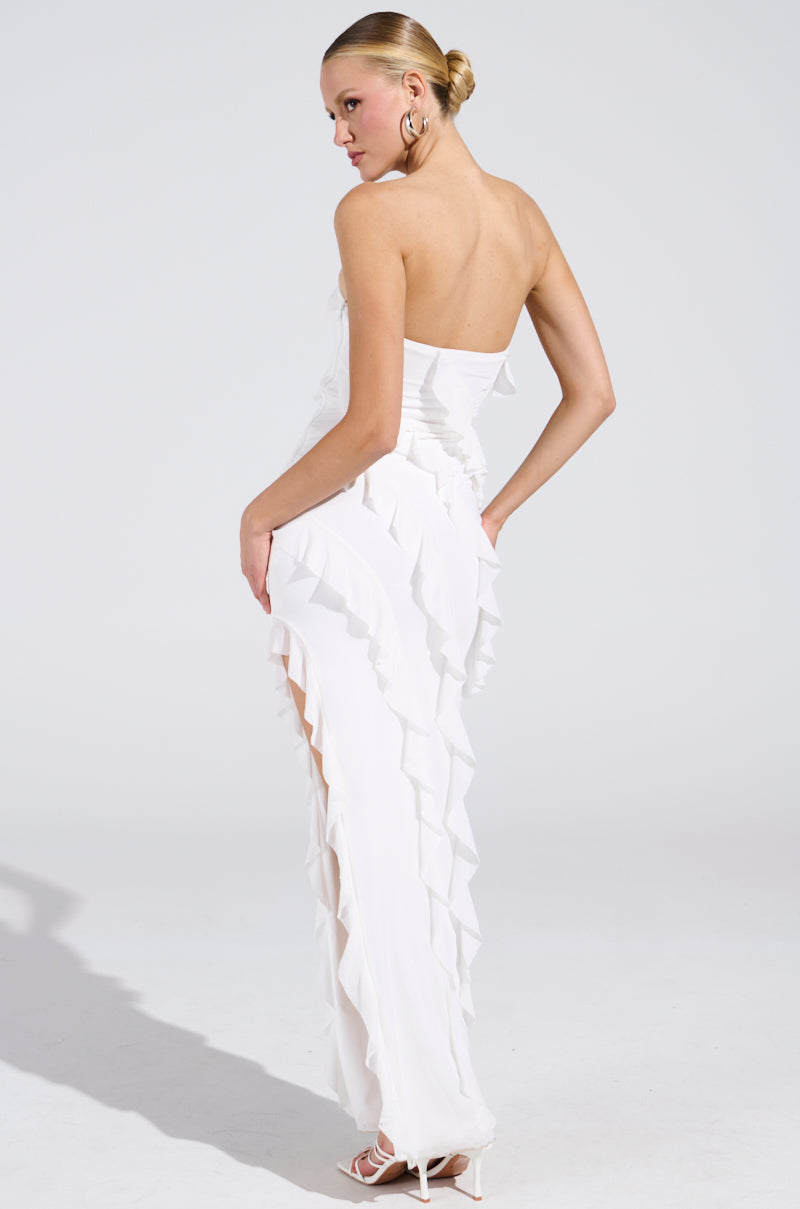A person models the ELLA MESH TUBE DRESS IN WHITE, a strapless piece with cascading ruffles. Styled in an elegant updo, they showcase large hoop earrings and high heels against a simple backdrop.