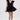 A woman in the ONCE UPON A DREAM WOVEN MINI DRESS IN BLACK, featuring a plunging V-neck, puffed shoulders, and bubble skirt, holds a silver clutch. She has slicked-back hair, statement earrings, and studded heeled sandals.