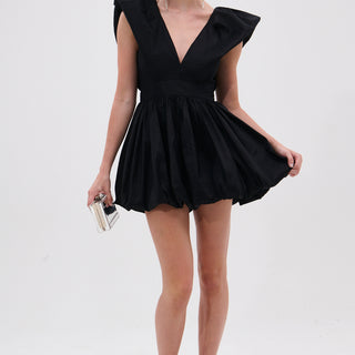 A woman in the ONCE UPON A DREAM WOVEN MINI DRESS IN BLACK, featuring a plunging V-neck, puffed shoulders, and bubble skirt, holds a silver clutch. She has slicked-back hair, statement earrings, and studded heeled sandals.