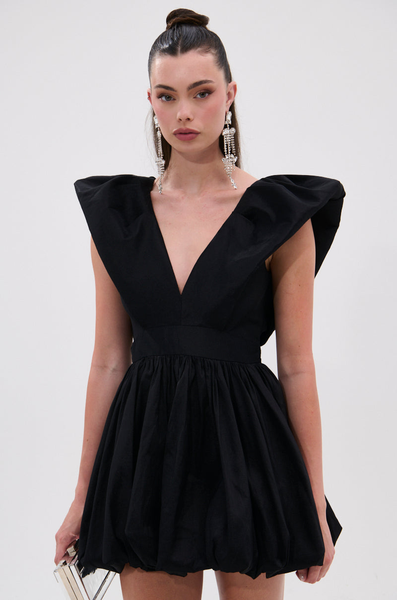 A woman models the ONCE UPON A DREAM WOVEN MINI DRESS IN BLACK with structured shoulders and a plunging V-neckline, accessorized with long earrings and a small clutch, her hair in a sleek high ponytail against a white background.