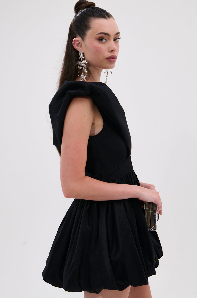 A woman poses sideways in the ONCE UPON A DREAM WOVEN MINI DRESS IN BLACK, styled with a small clutch and statement earrings. Her long, dark hair is half-up in a top knot, set against a plain, light background.