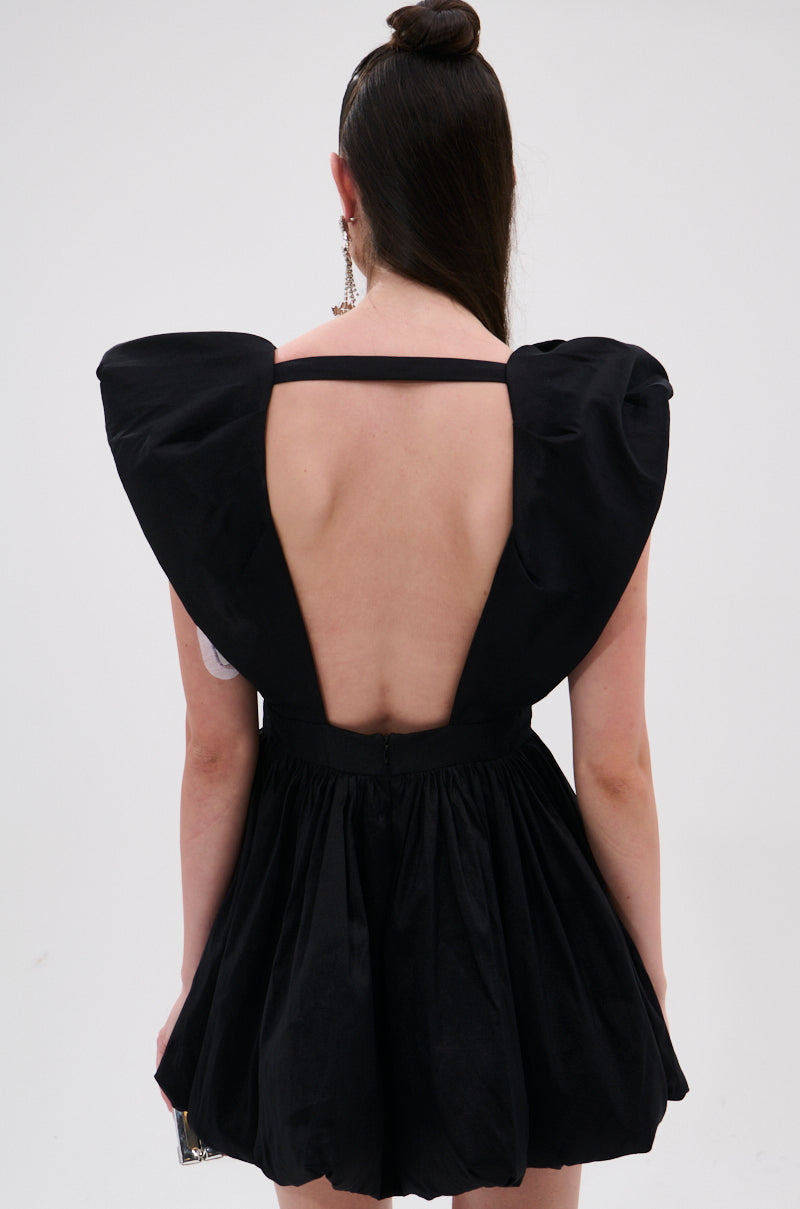A woman with long dark hair in a top bun is seen from behind wearing the ONCE UPON A DREAM WOVEN MINI DRESS IN BLACK, featuring large puffed shoulders, an open back, and a chic skater silhouette against a plain white background.