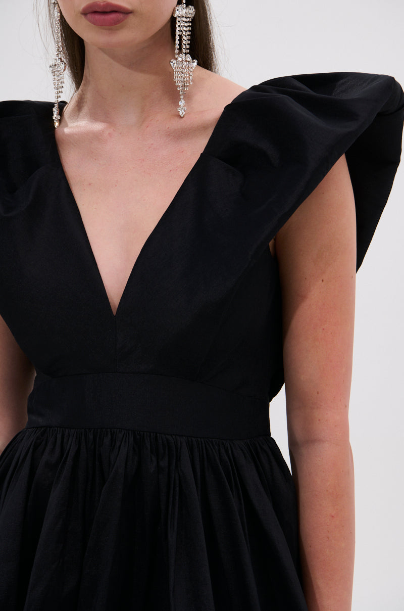 A woman models the ONCE UPON A DREAM WOVEN MINI DRESS IN BLACK with structured shoulders and a plunging V-neckline; she wears long pearl earrings and is shown from shoulders to mid-torso against a plain background.