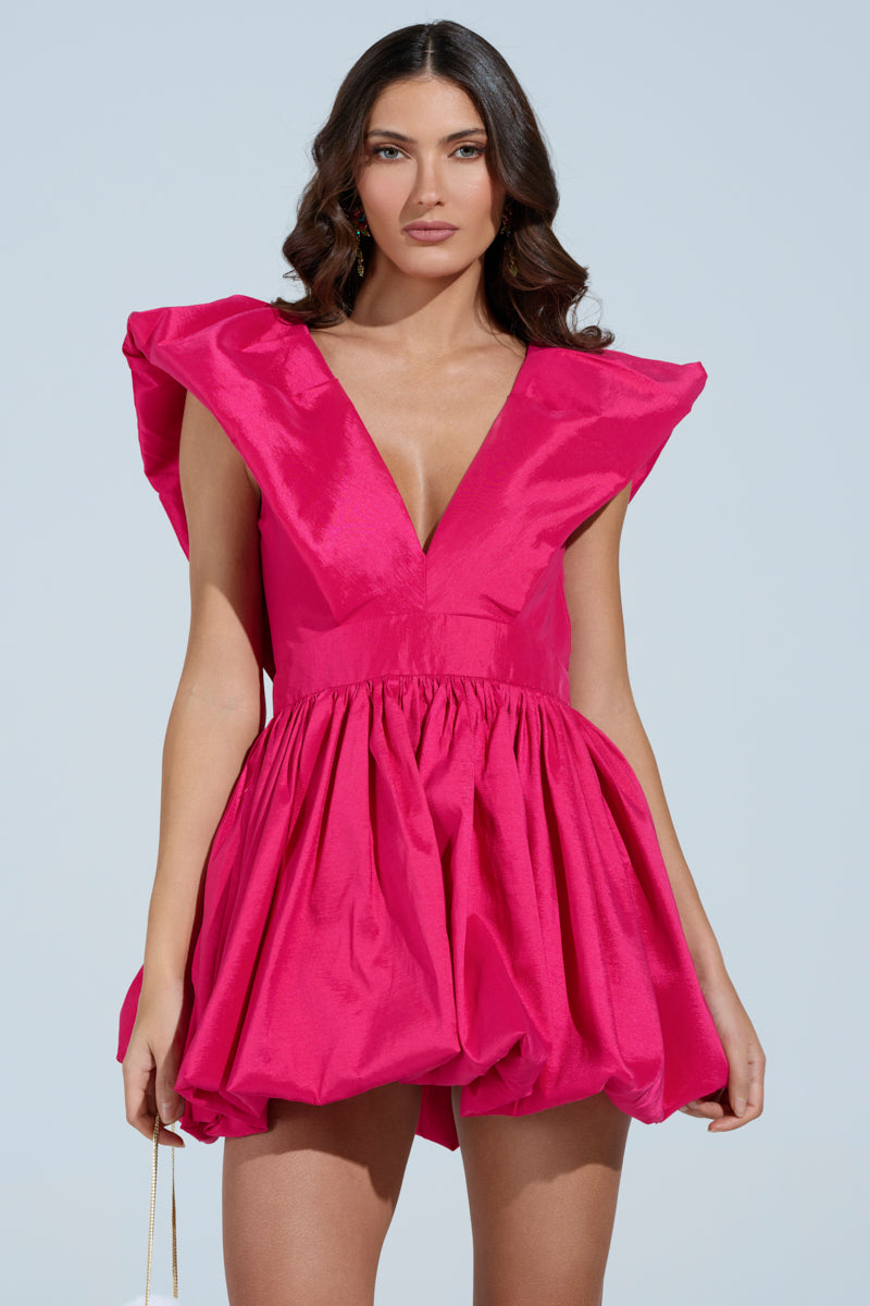 A woman in the ONCE UPON A DREAM WOVEN MINI DRESS, a vibrant pink mini dress with a plunging V-neckline, skater silhouette, and pronounced shoulders poses against a light blue background. She has long, wavy hair and hoop earrings, exuding effortless elegance. 1