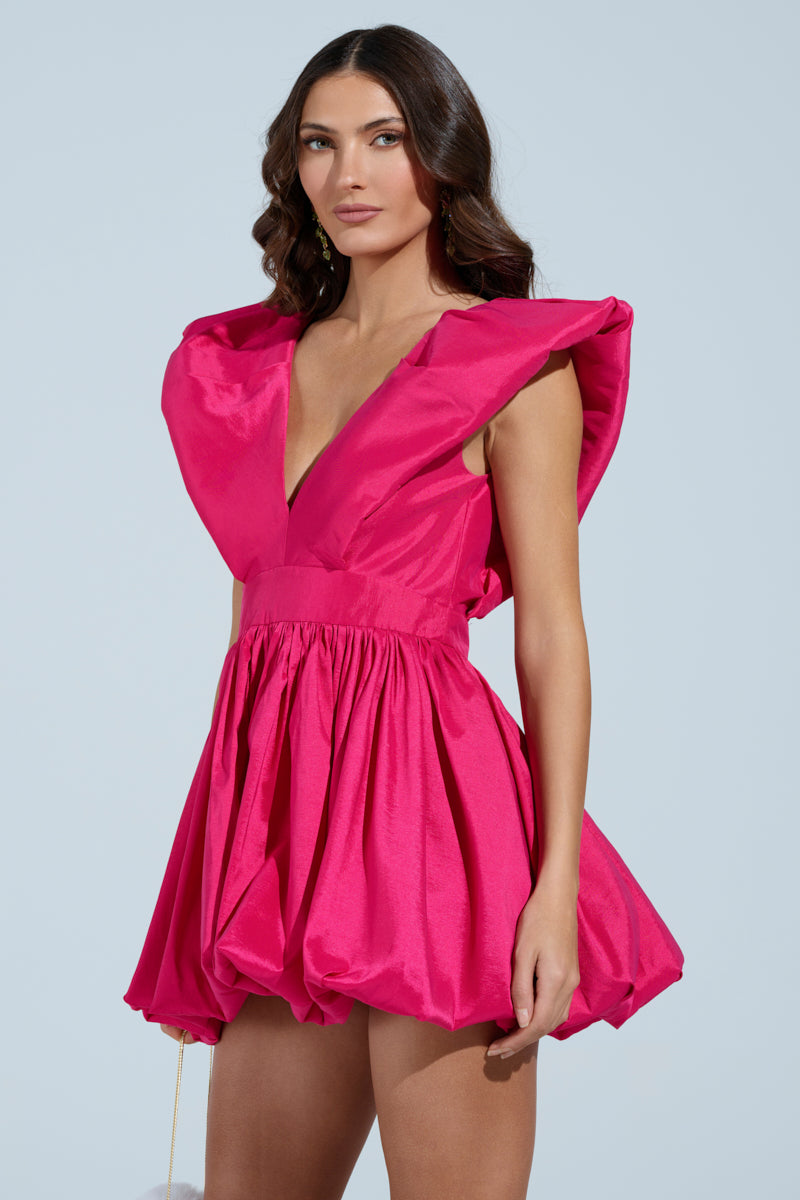 In a bright pink ONCE UPON A DREAM WOVEN MINI DRESS with dramatic shoulders, skater silhouette, plunging V-neckline, and cinched waist, a woman stands confidently. Her long wavy hair cascades against the neutral backdrop as she wears the dress's short balloon-like skirt. 3