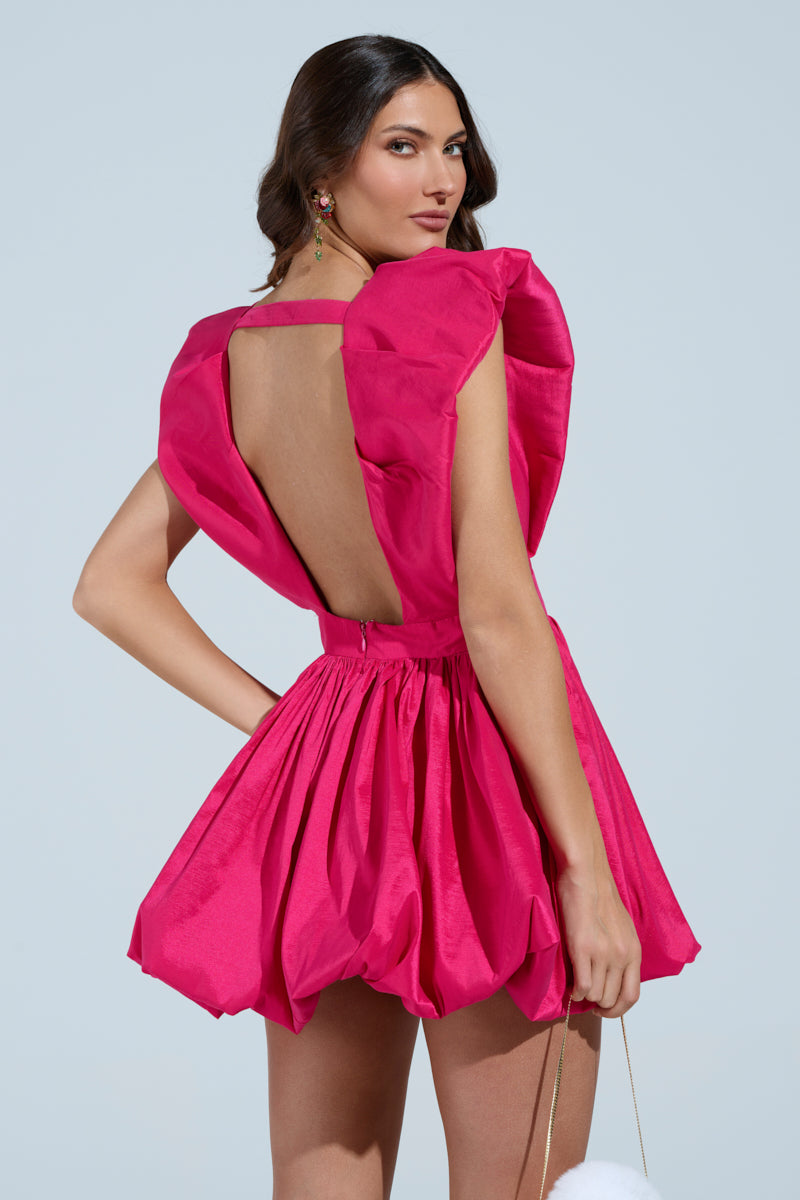 A woman poses with her back turned, wearing the ONCE UPON A DREAM WOVEN MINI DRESS in vibrant pink, featuring an open back and ruffled shoulders. The dress has a short, pleated skater silhouette with a cinched waist. She holds a white fuzzy accessory in one hand against a light background. 5