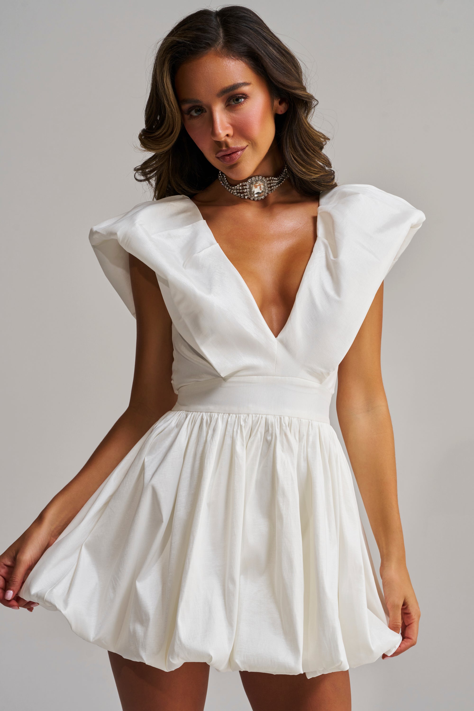 A woman poses against a plain background in the FAR GONE WHITE MINI DRESS, a skater-style mini dress with puffed shoulders, a fitted waist, and deep V-neck, paired with a chunky pearl choker necklace.
