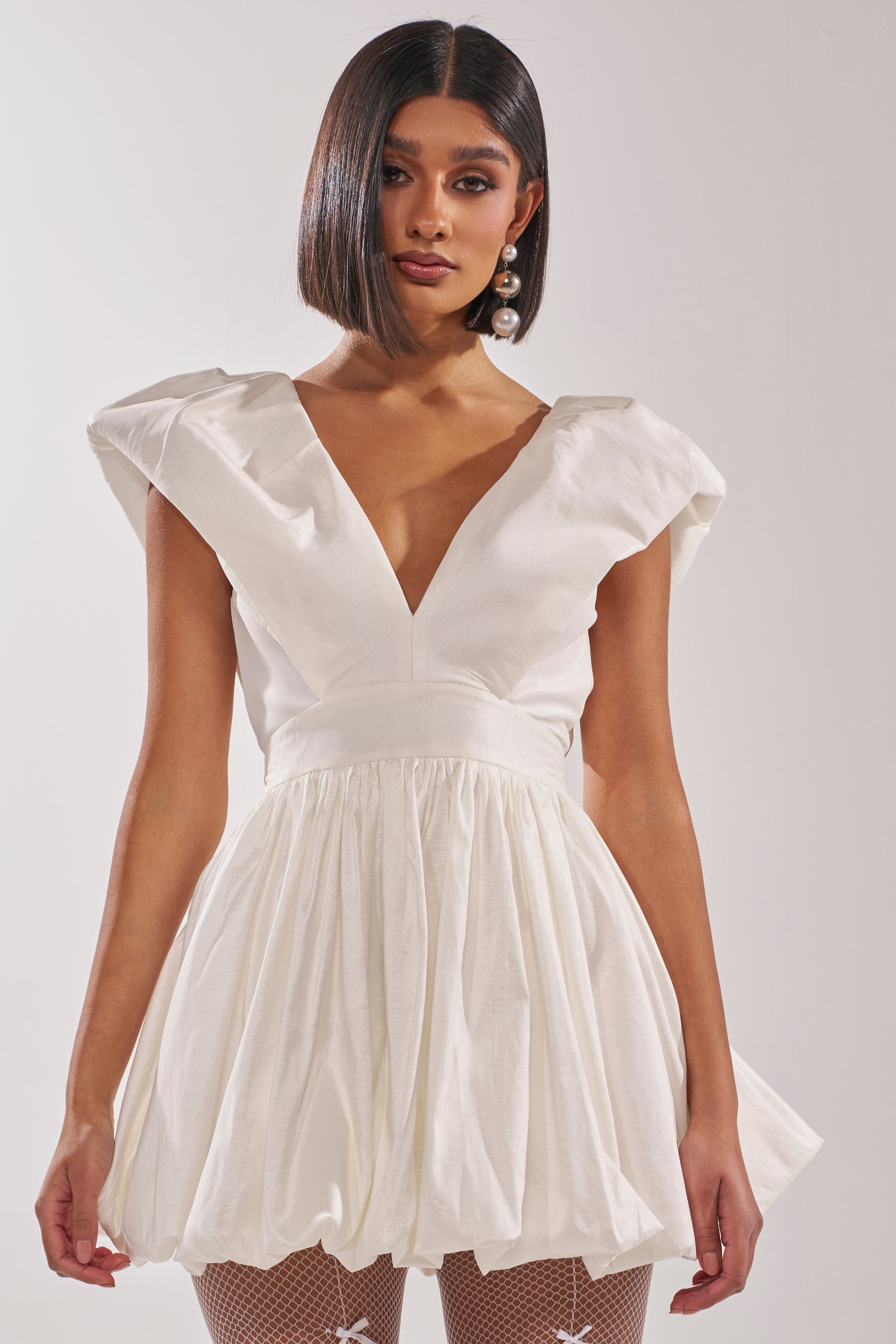 A woman with a sleek bob wears the FAR GONE WHITE MINI DRESS, styled with puff sleeves, a plunging V-neckline, and a skater silhouette. She pairs it with dangling pearl earrings and poses against a plain background.