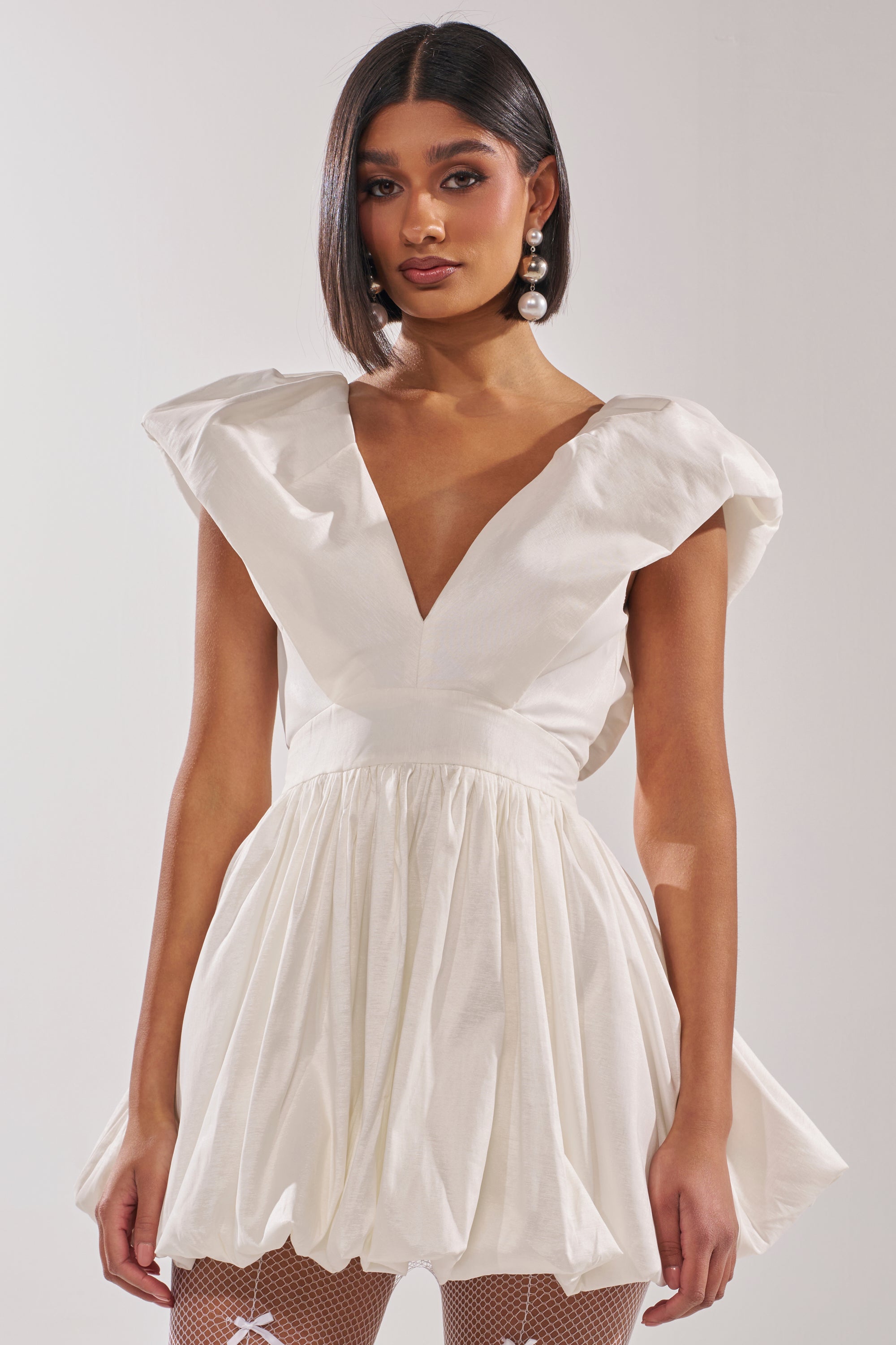 A woman with a sleek bob wears the FAR GONE WHITE MINI DRESS, featuring puffed sleeves and a plunging V-neckline. She accessorizes with large pearl earrings and stands against a plain light background.
