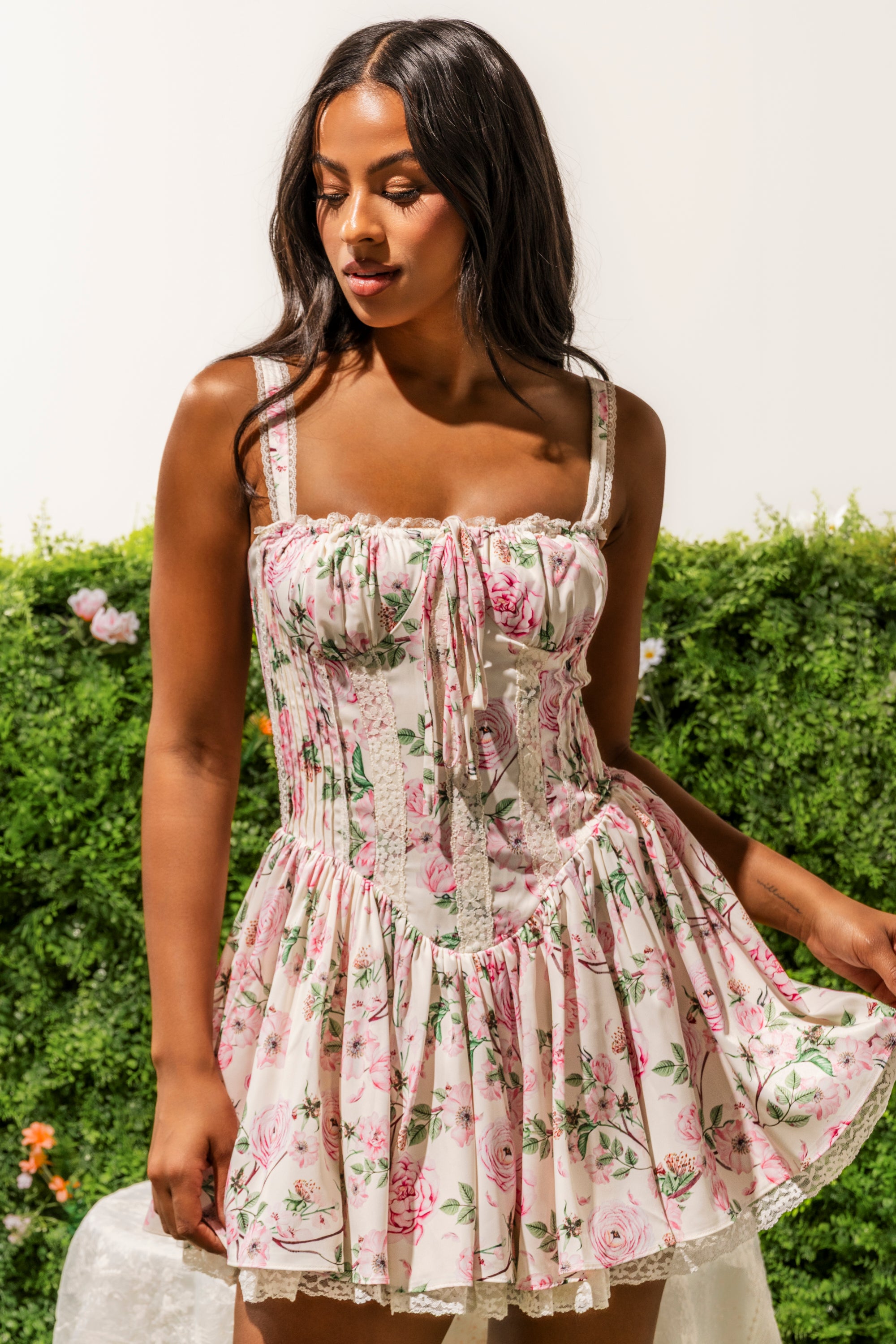 A woman in the SELENE FLORAL MINI DRESS stands outdoors, gently holding the skirt of her pink floral fit-and-flare mini dress. Surrounded by green foliage and flowers, she looks down as her long wavy hair cascades over her shoulder. 1