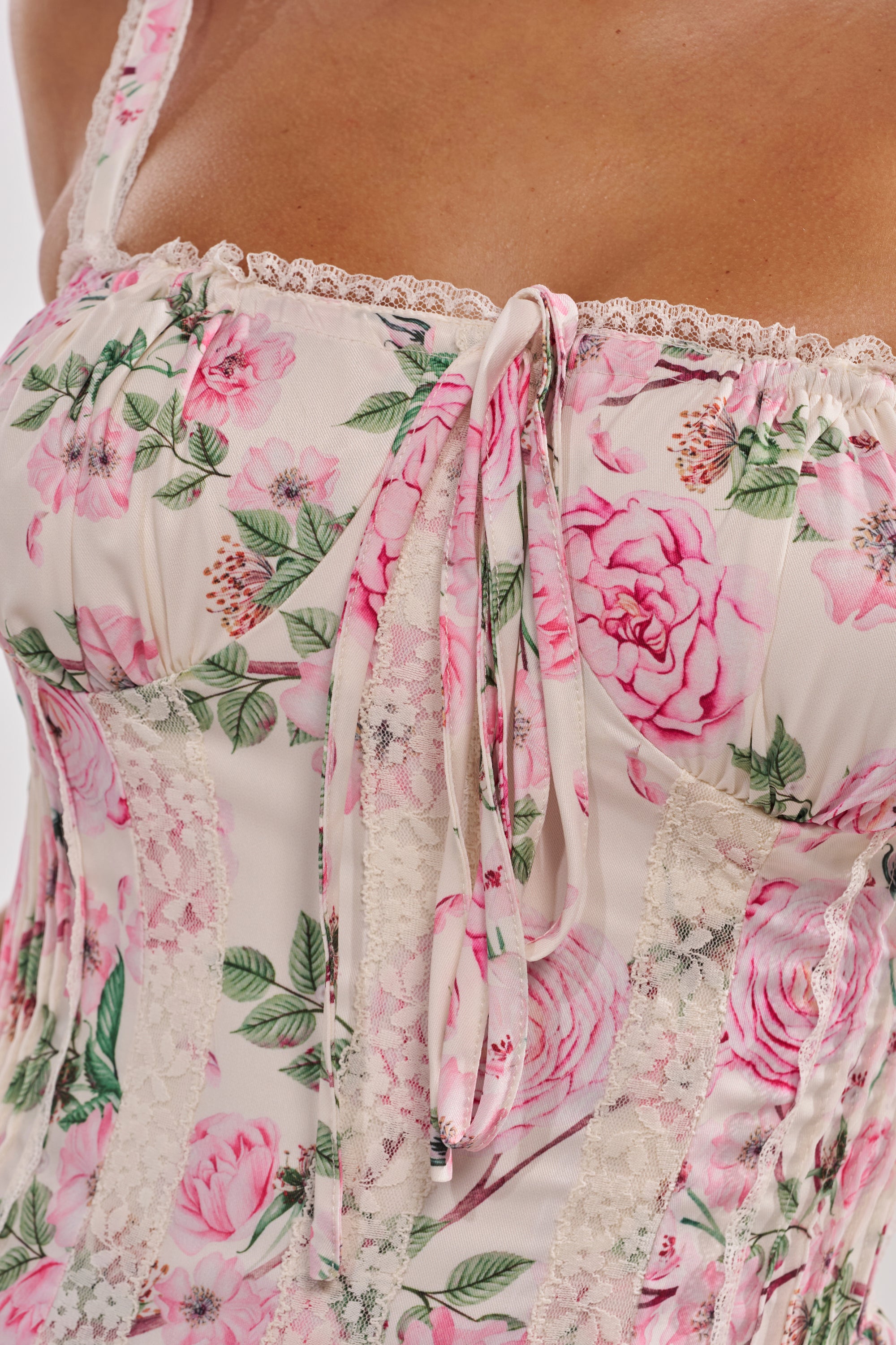 Close-up of a person wearing the SELENE FLORAL MINI DRESS, featuring pink and white fabric with large roses, green leaves, lace trim, and a bow at the neckline—perfect for spring. 7