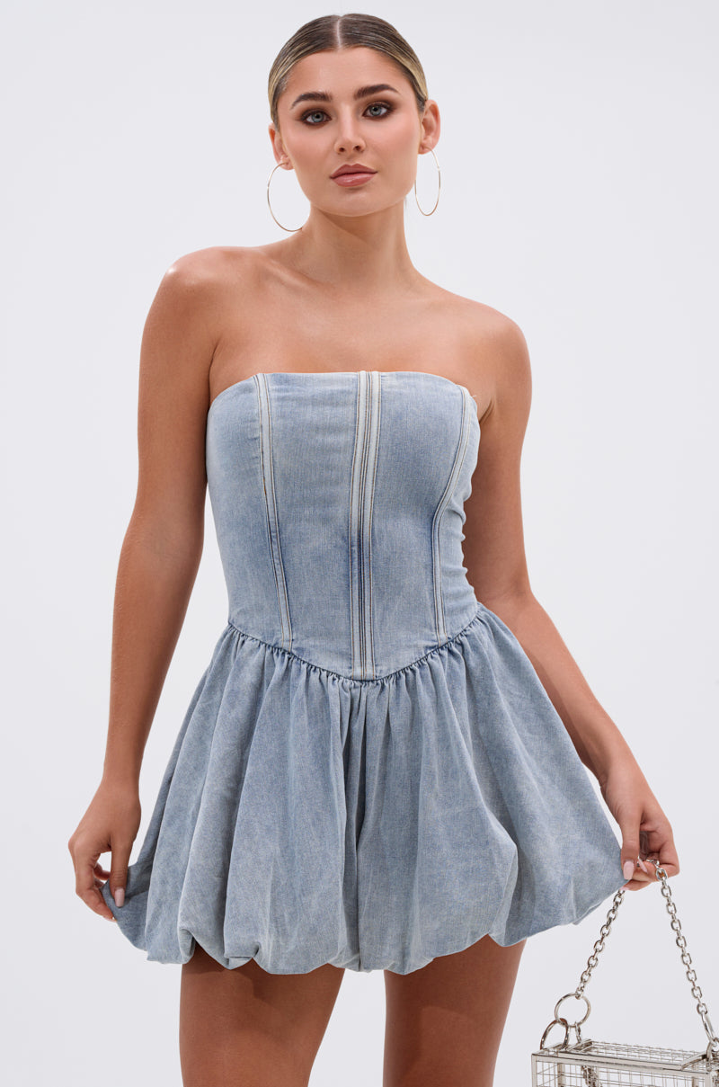 She wears the BELLA LOO MINI DRESS IN DENIM, featuring a strapless corset bodice and flared bubble skirt. Her look is finished with hoop earrings, a silver bag, and chic pulled-back hair. 0