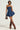 A woman confidently poses in the BELLA LOO NAVY MINI DRESS, featuring a strapless neckline. She carries a small brown handbag and pairs the look with knee-high brown boots. Her short hair is styled neatly, enhanced by tasteful jewelry.