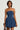 Wearing the BELLA LOO NAVY MINI DRESS, a woman with short hair holds a small handbag. Her outfit features a strapless design with a flared skirt. She accessorizes with a cross necklace and small earrings, all set against a plain, light-colored background that complements her sophisticated appearance.