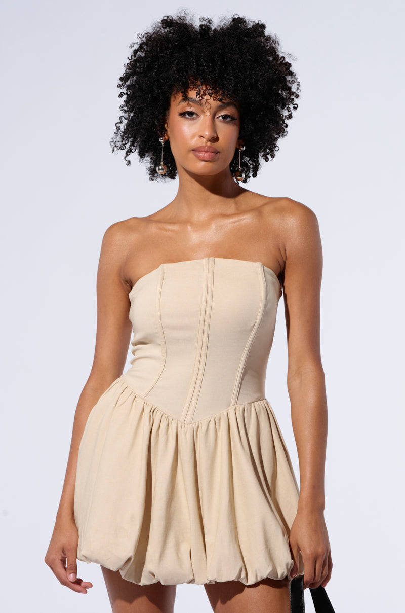 A person with curly hair poses against a plain background, wearing a strapless, beige BELLA LOO MINI DRESS that features a skater dress silhouette and non-stretch knit fabric. The individual's expression is neutral, with both arms relaxed by their sides.