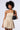 A person with curly hair stands against a plain background, wearing the BELLA LOO MINI DRESS in a beige, strapless, bubble-hem silhouette. They hold a small, black shoulder bag with a glossy finish and wear small hoop earrings.