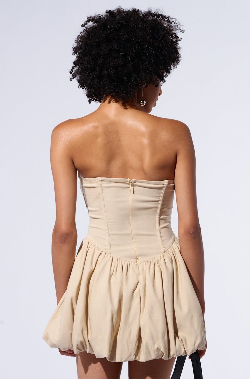 A person with short, curly black hair is pictured from the back wearing the BELLA LOO MINI DRESS. The cream-colored, strapless skater dress showcases non-stretch knit fabric with a fitted bodice and a gathered voluminous skirt, complemented by a visible zipper down the back. The background is plain and light-colored.