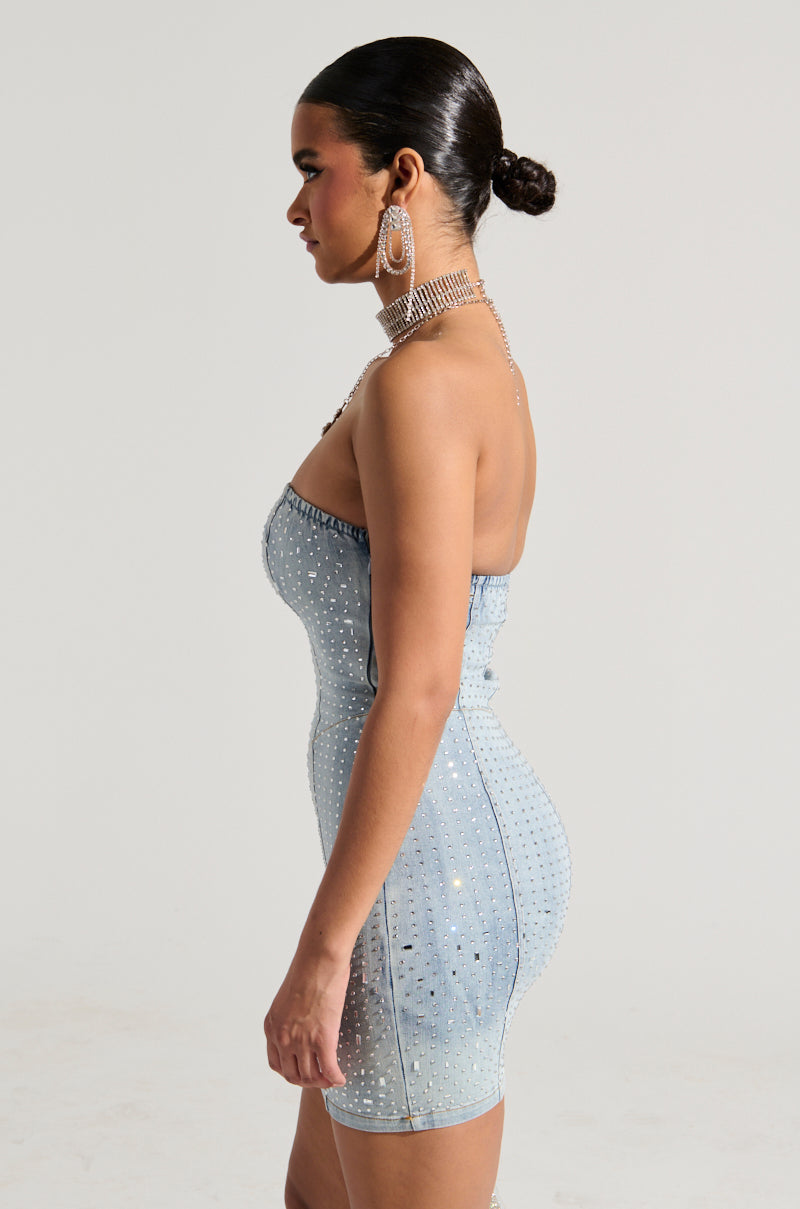 A woman wearing the JUST WANNA ROCK Denim Rhinestone Mini Dress stands sideways, showcasing its strapless design. She accessorizes with large hoop earrings and has styled her hair in a sleek bun. The dress's shimmering rhinestone embellishments catch the light against the plain background. 3