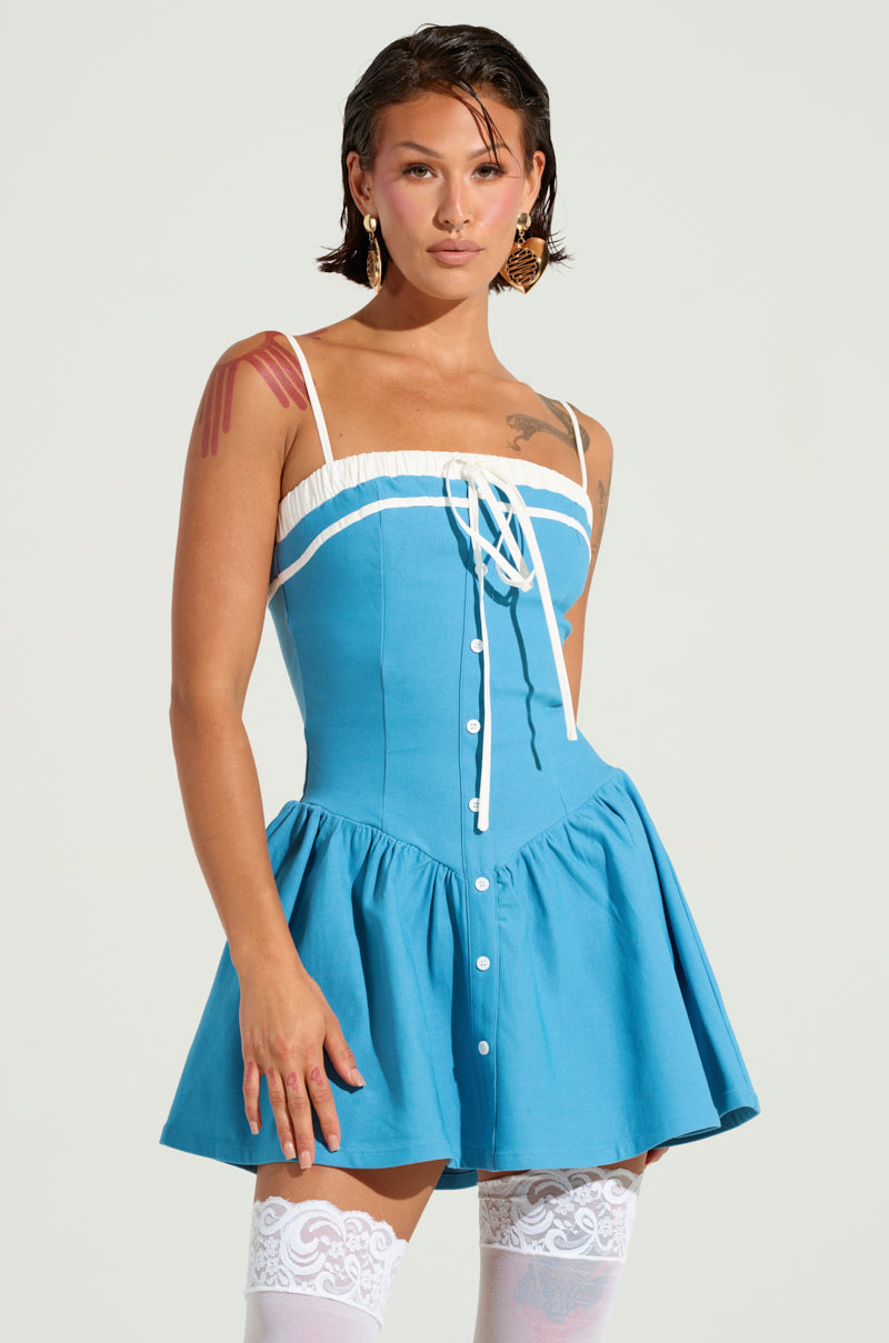 A person wearing the TOO SWEET POPLIN MINI DRESS, featuring spaghetti straps and adorned with white buttons, stands against a plain background. The dress boasts a corset-inspired bodice and is paired with tattoos on their arm and chest, lace-topped stockings, and large earrings.