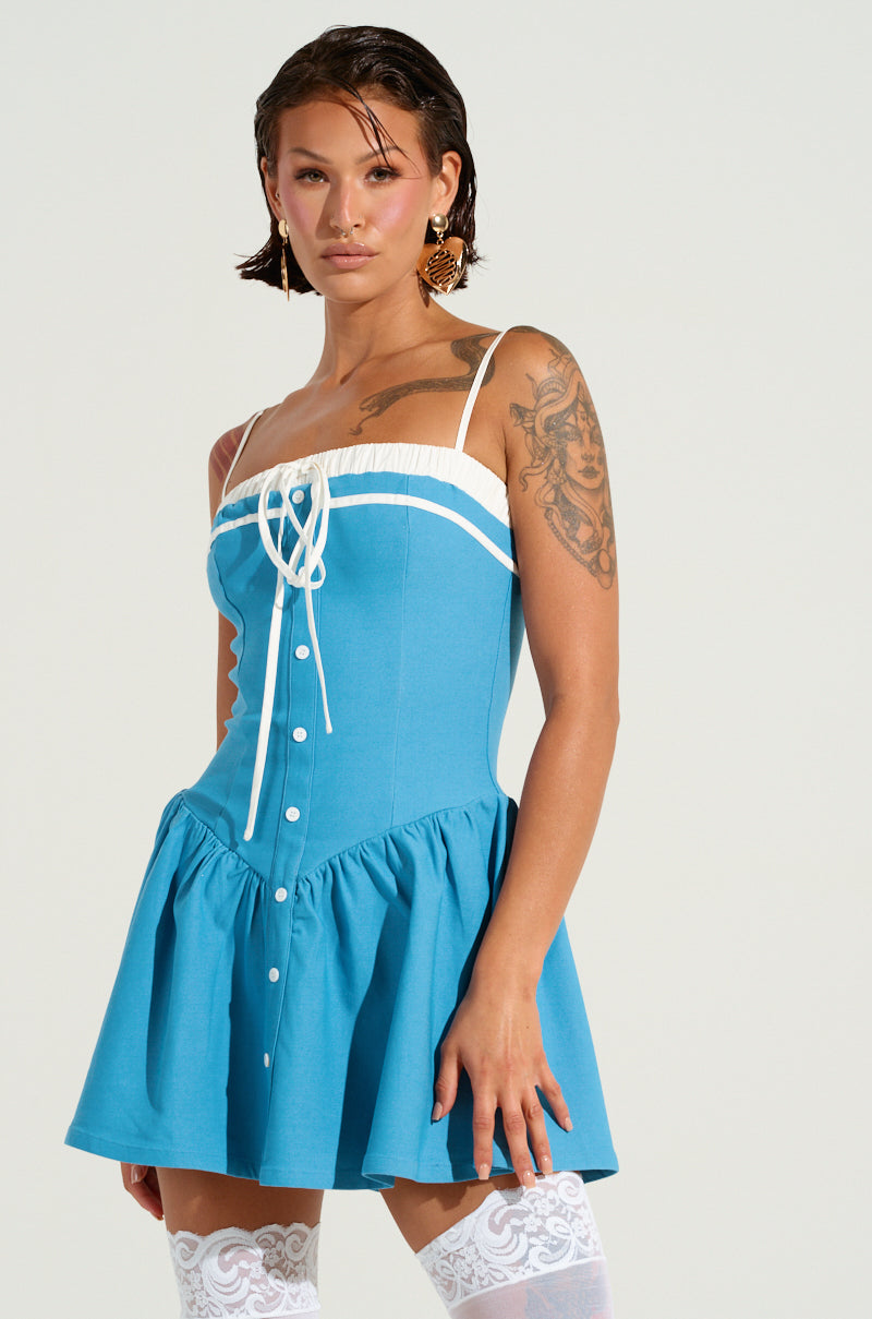 A person confidently stands wearing the TOO SWEET POPLIN MINI DRESS, a sleeveless blue mini featuring white trim and buttons. Its corset-inspired bodice and flared skirt beautifully complement their short, slicked-back hair and visible tattoos, while sheer white thigh-high stockings complete the look.