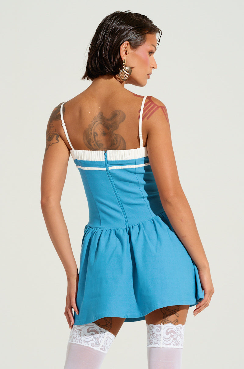 A person with tattooed arms and shoulder-length dark hair stands facing away, wearing the TOO SWEET POPLIN MINI DRESS, which features a blue fabric with white straps and a ruffled skirt. The dress has a corset-inspired bodice and is complemented by their white lace thigh-high stockings.