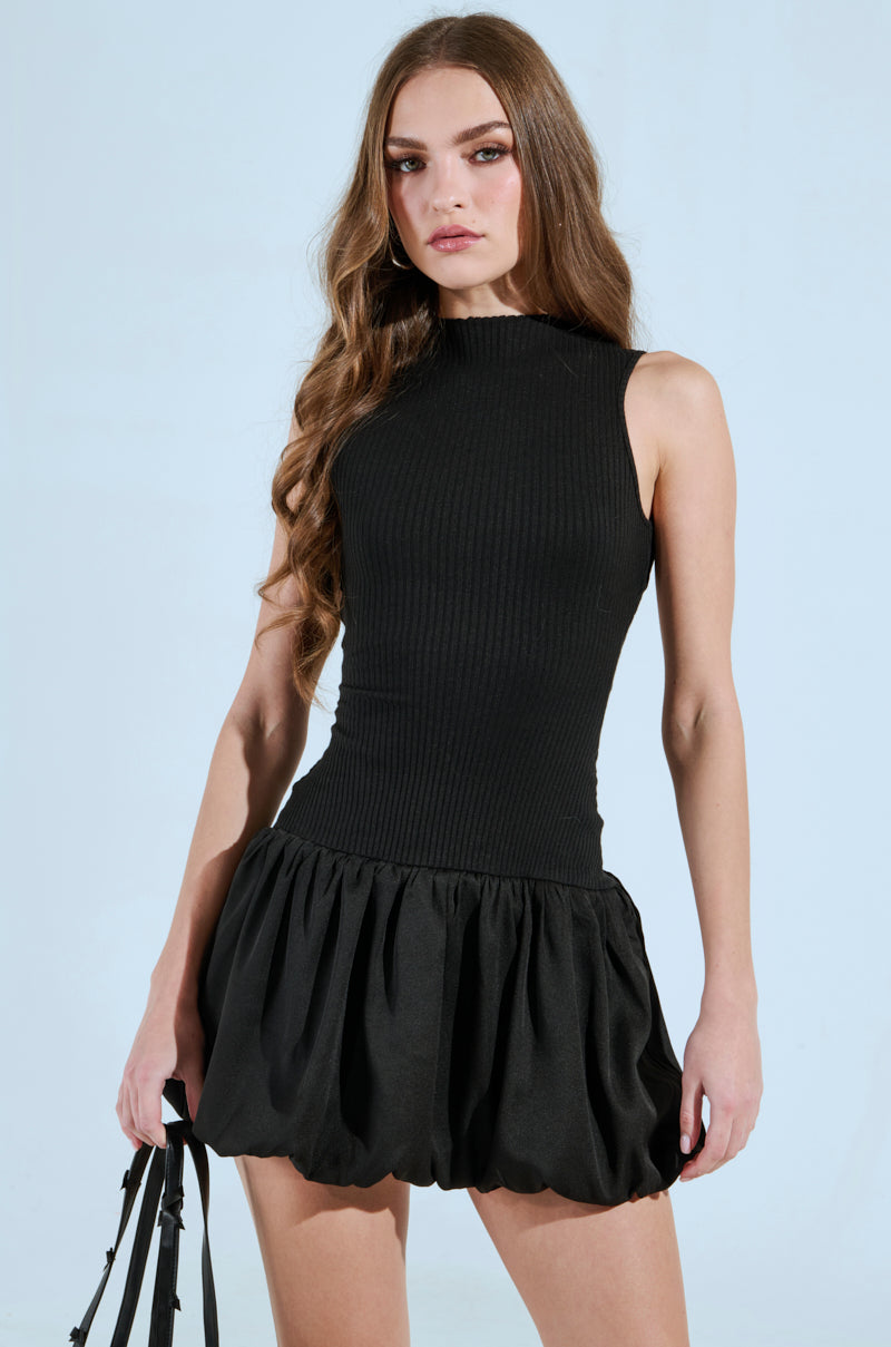 A person with long hair wears the DOWN FOR WHATEVER MINI DRESS, a sleeveless black outfit with a fitted ribbed knit top, voluminous skirt, and chic back cut-out. They hold a strapped black accessory against a plain light background.