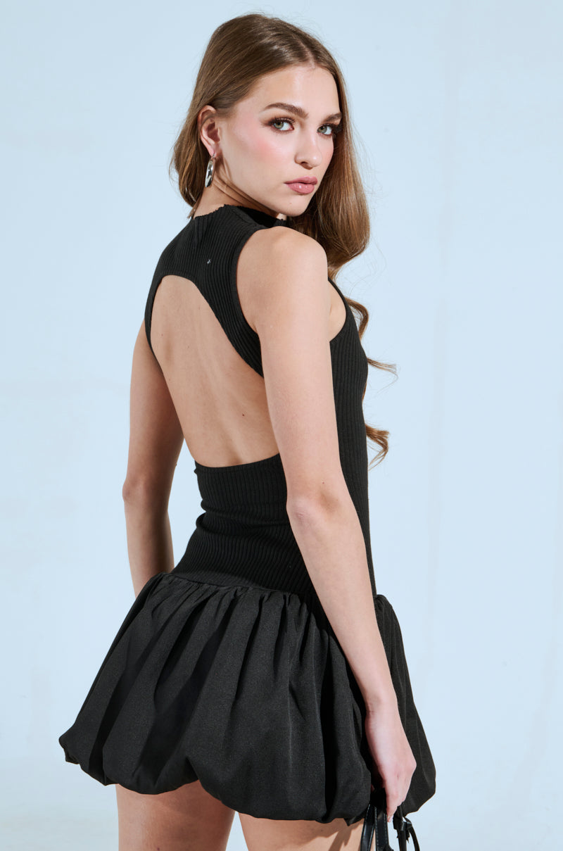 A person with long, wavy hair wears the "DOWN FOR WHATEVER MINI DRESS," a sleeveless black dress featuring a dramatic back cut-out and voluminous short skirt. They look over their shoulder against a light blue background.