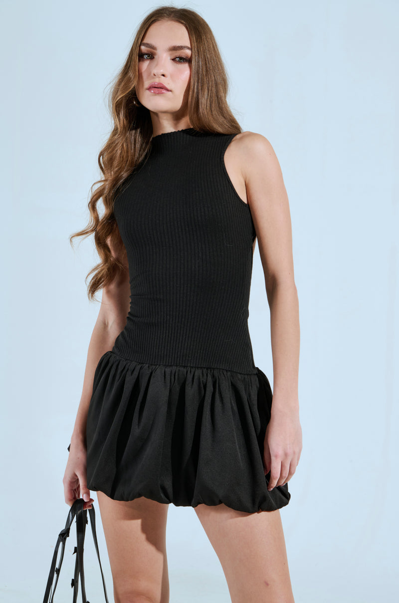 A woman with long hair in a "Down For Whatever Mini Dress" stands against a light blue background, her black bag enhancing the grace of her ensemble.