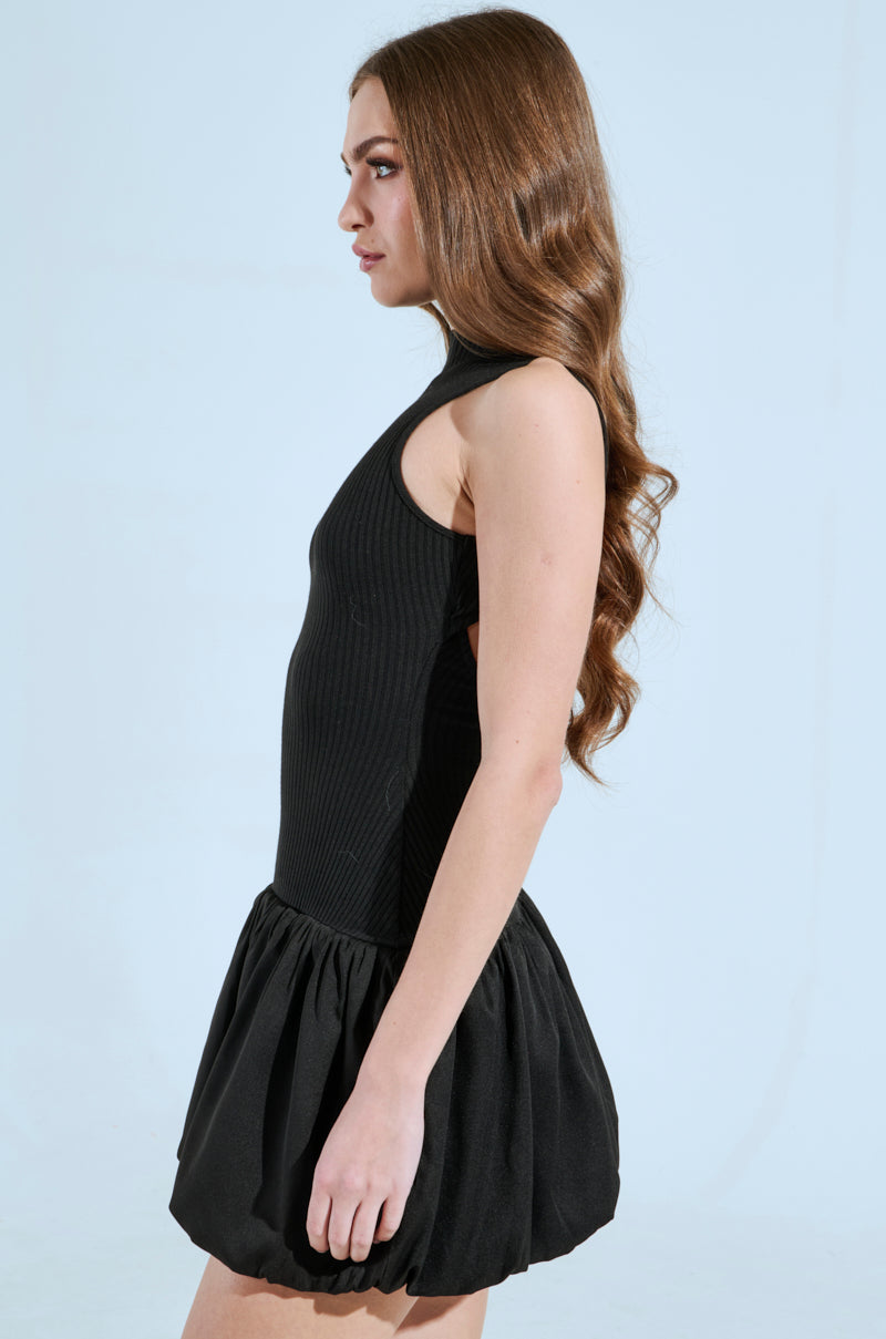 A woman with long wavy hair stands in profile against a light background, wearing the sleeveless "DOWN FOR WHATEVER MINI DRESS" featuring a ribbed knit fabric that complements her figure and a short, voluminous skirt. A subtle back cut-out enhances its chic appeal.