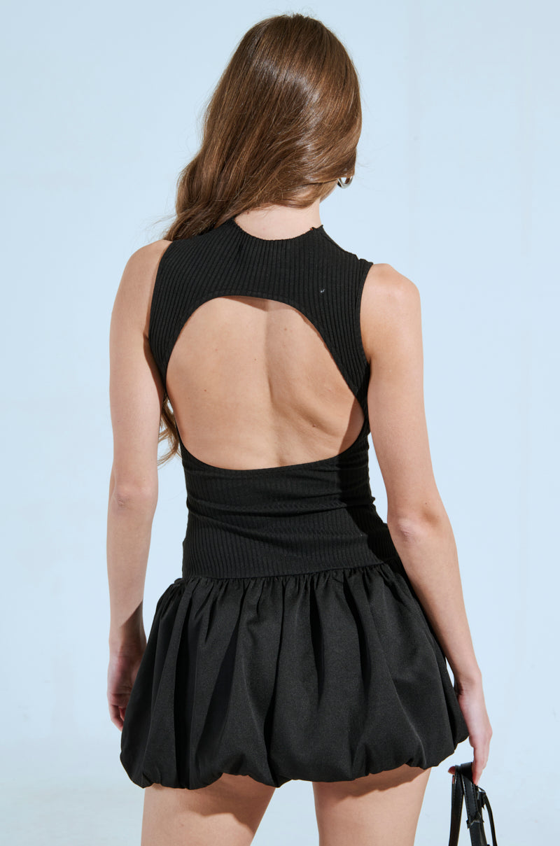 A woman with long hair is seen from behind wearing the DOWN FOR WHATEVER MINI DRESS, a sleeveless black mini dress with a chic back cut-out and puffy skirt. She holds a black bag in her left hand against a plain, light-colored background.
