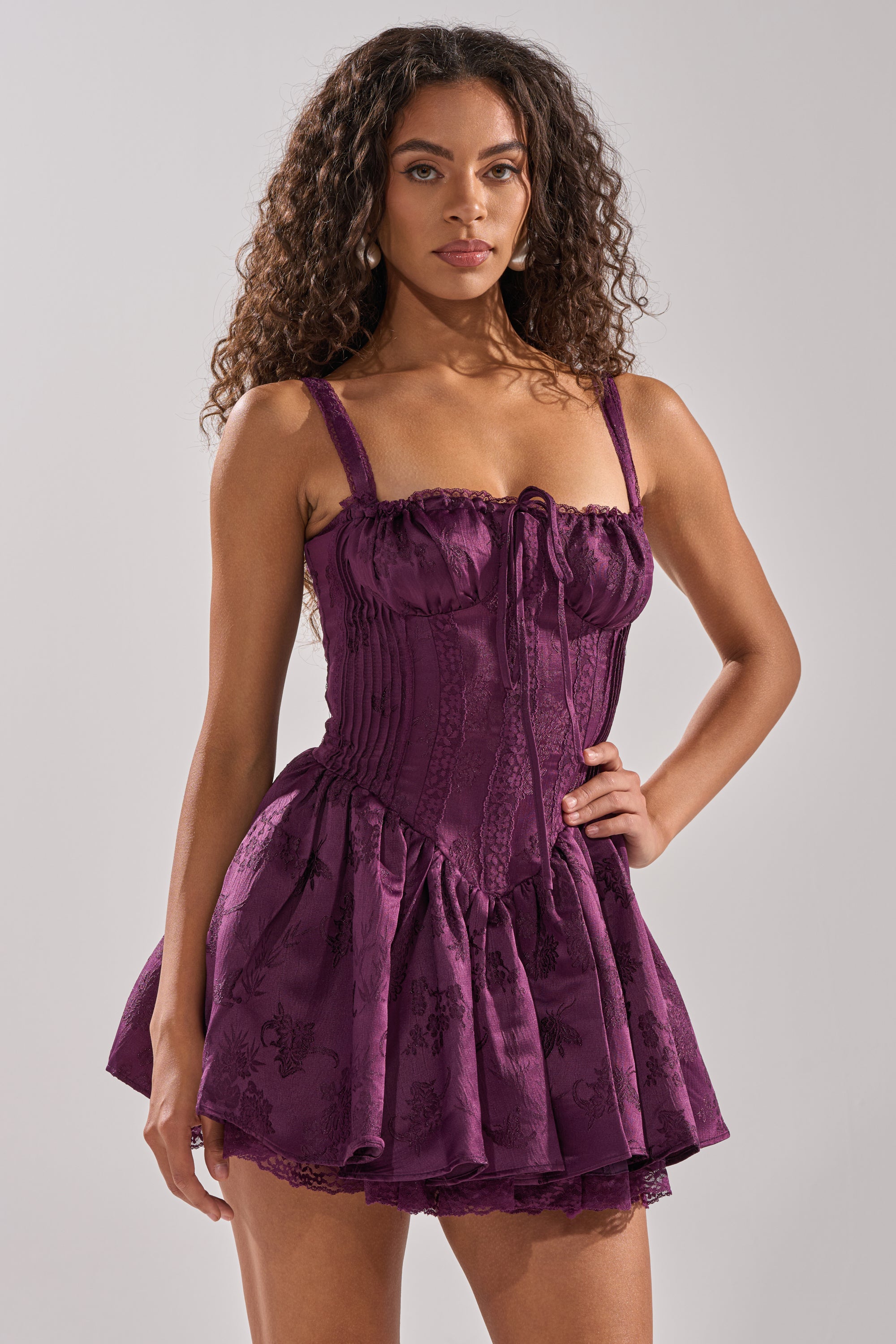 A woman with long curly hair models the DANCE WITH ME MINI DRESS IN PLUM, a sleeveless satin jacquard mini with a corset-style bodice and lace details, standing confidently against a neutral background. 0