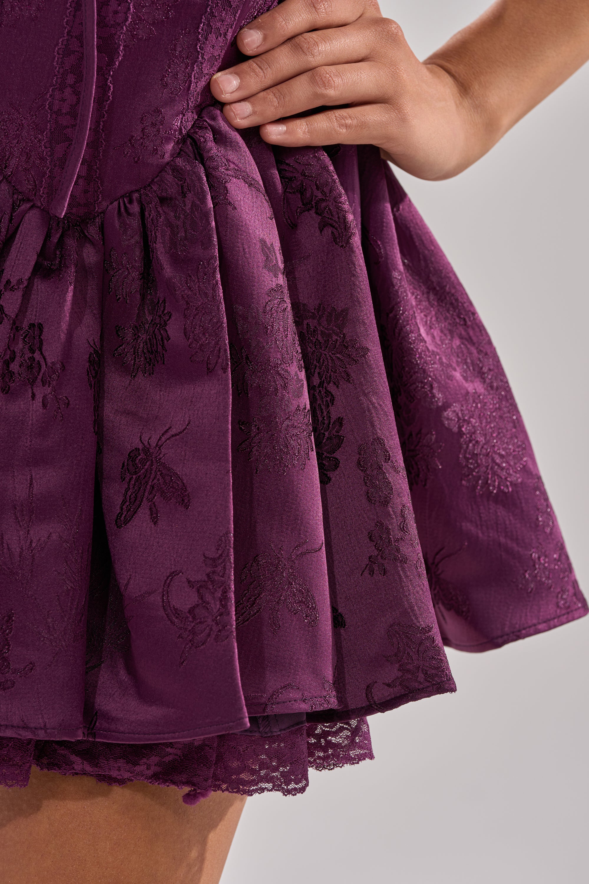 A close-up of a person wearing the DANCE WITH ME MINI DRESS IN PLUM, featuring deep purple textured jacquard, floral patterns, a corset-style bodice, layered satin details, and a delicate lace finish. Their hand rests on their waist. 9