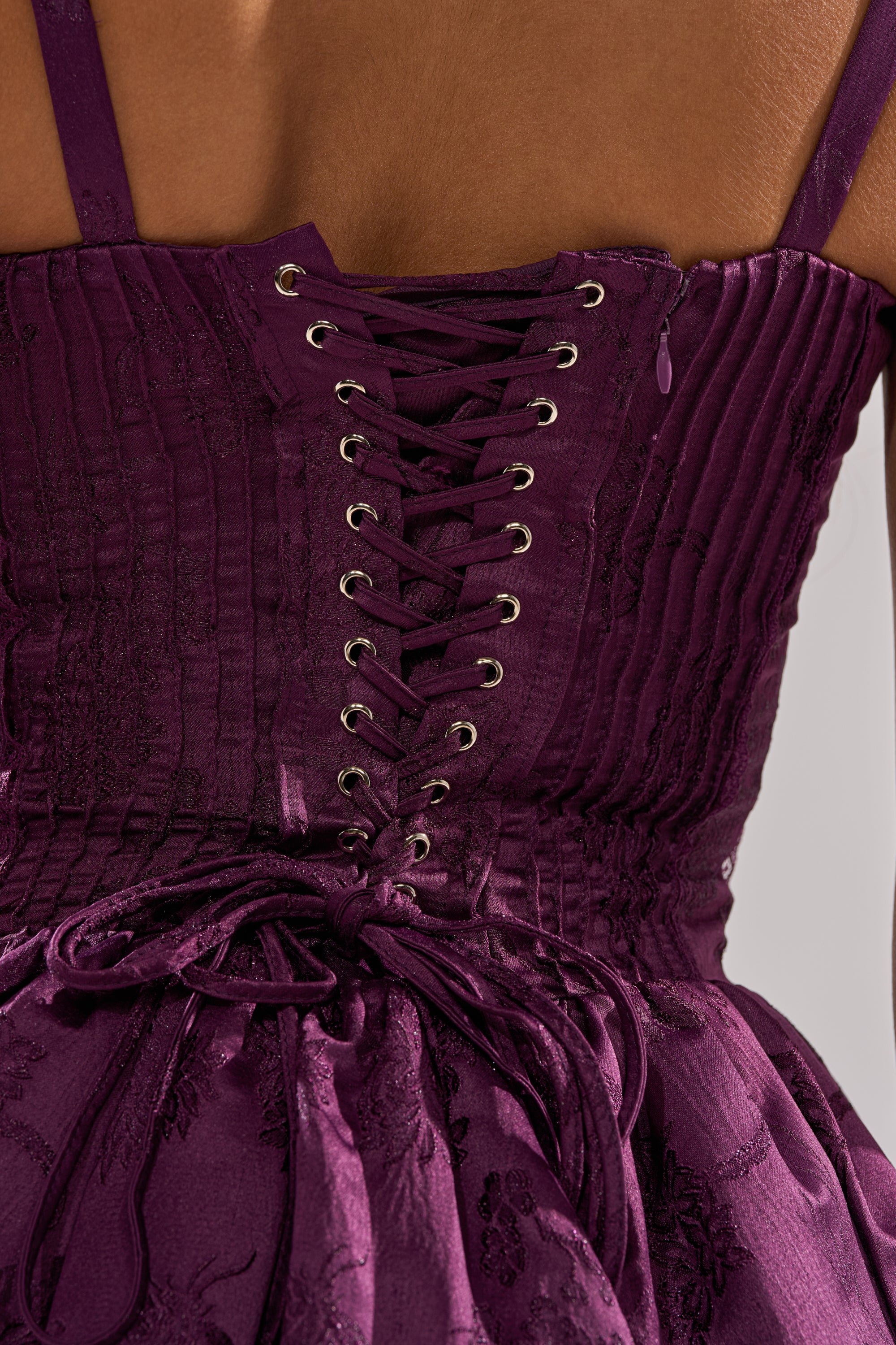 Close-up of a person wearing the DANCE WITH ME MINI DRESS IN PLUM, featuring deep purple satin jacquard fabric with a floral pattern, a corset-style bodice, intricate lace-up back with silver eyelets, and satin ribbon ties. 10