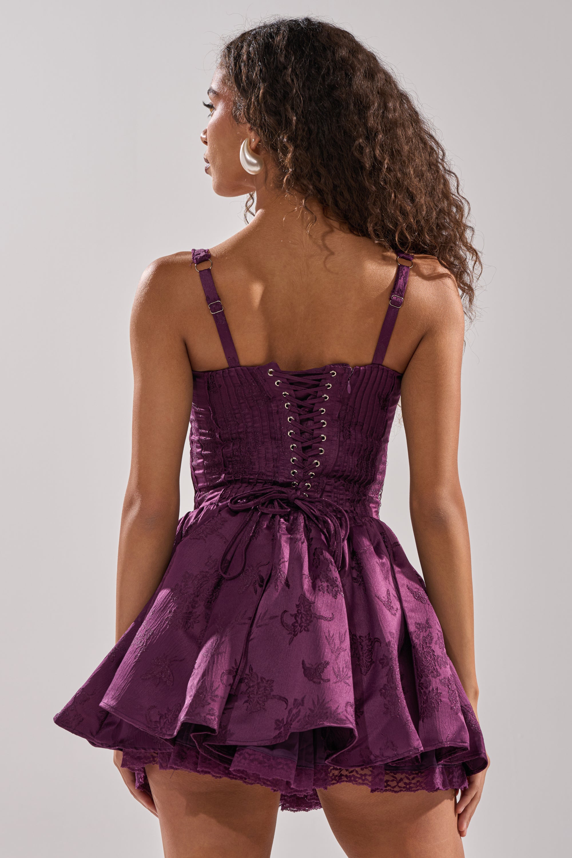 A woman with long curly hair, wearing the DANCE WITH ME MINI DRESS IN PLUM—featuring a lace-up corset bodice and flared skirt—stands facing away from the camera against a plain light background. 5