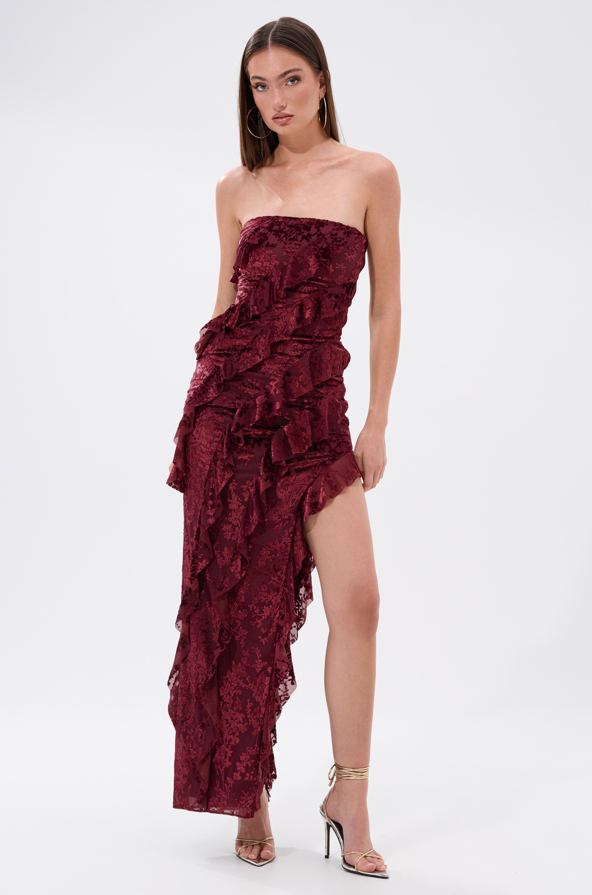 A woman models the WHERE WE GOIN VELVET BURNOUT MIDI DRESS, a strapless burgundy ruffled gown with a thigh-high slit, paired with gold high-heeled sandals, against a plain white background.
