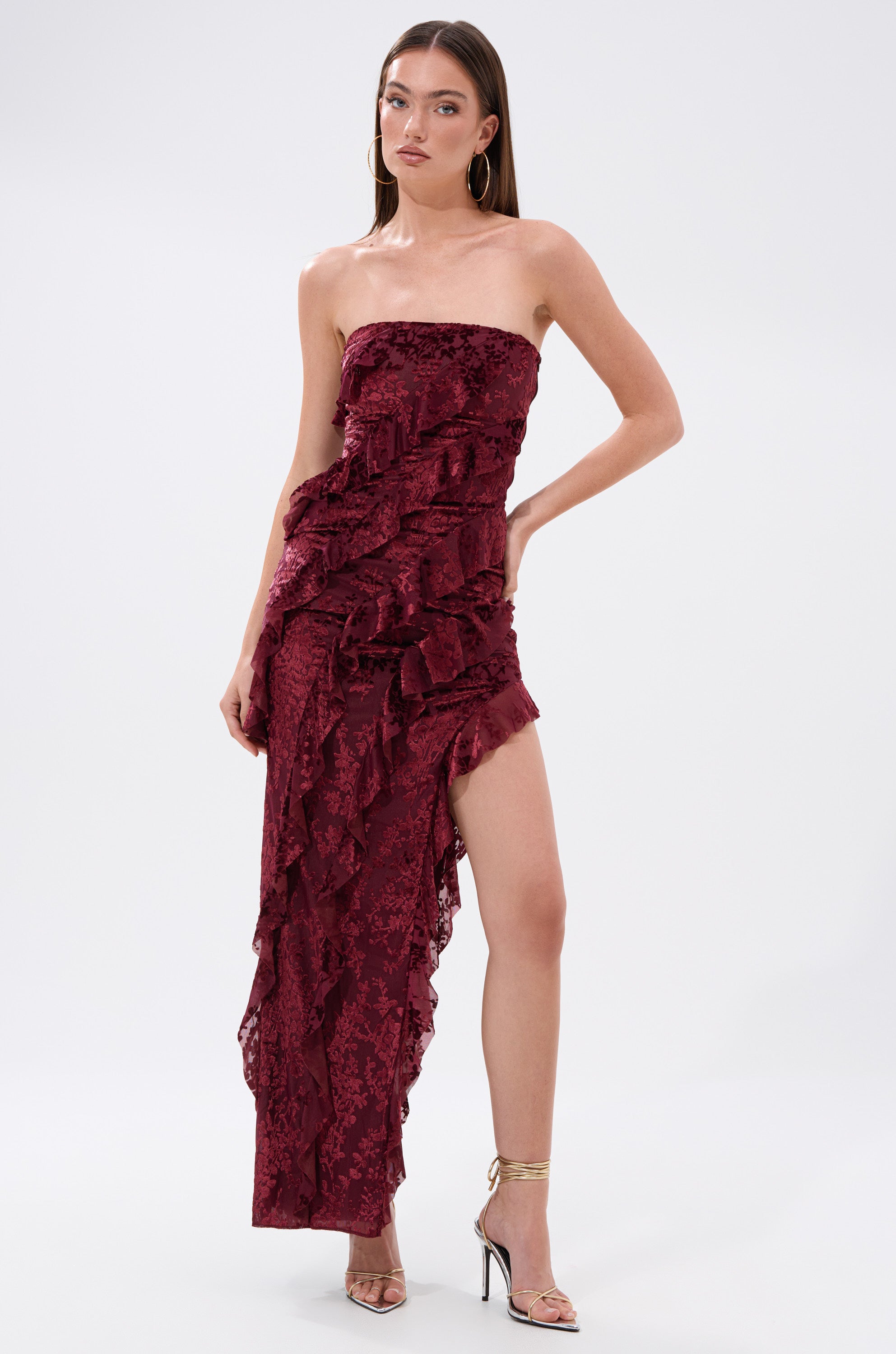 A woman models the WHERE WE GOIN VELVET BURNOUT MIDI DRESS—strapless, burgundy, with ruffles and a high slit—styled with gold heels and hoop earrings, posing against a plain white background.