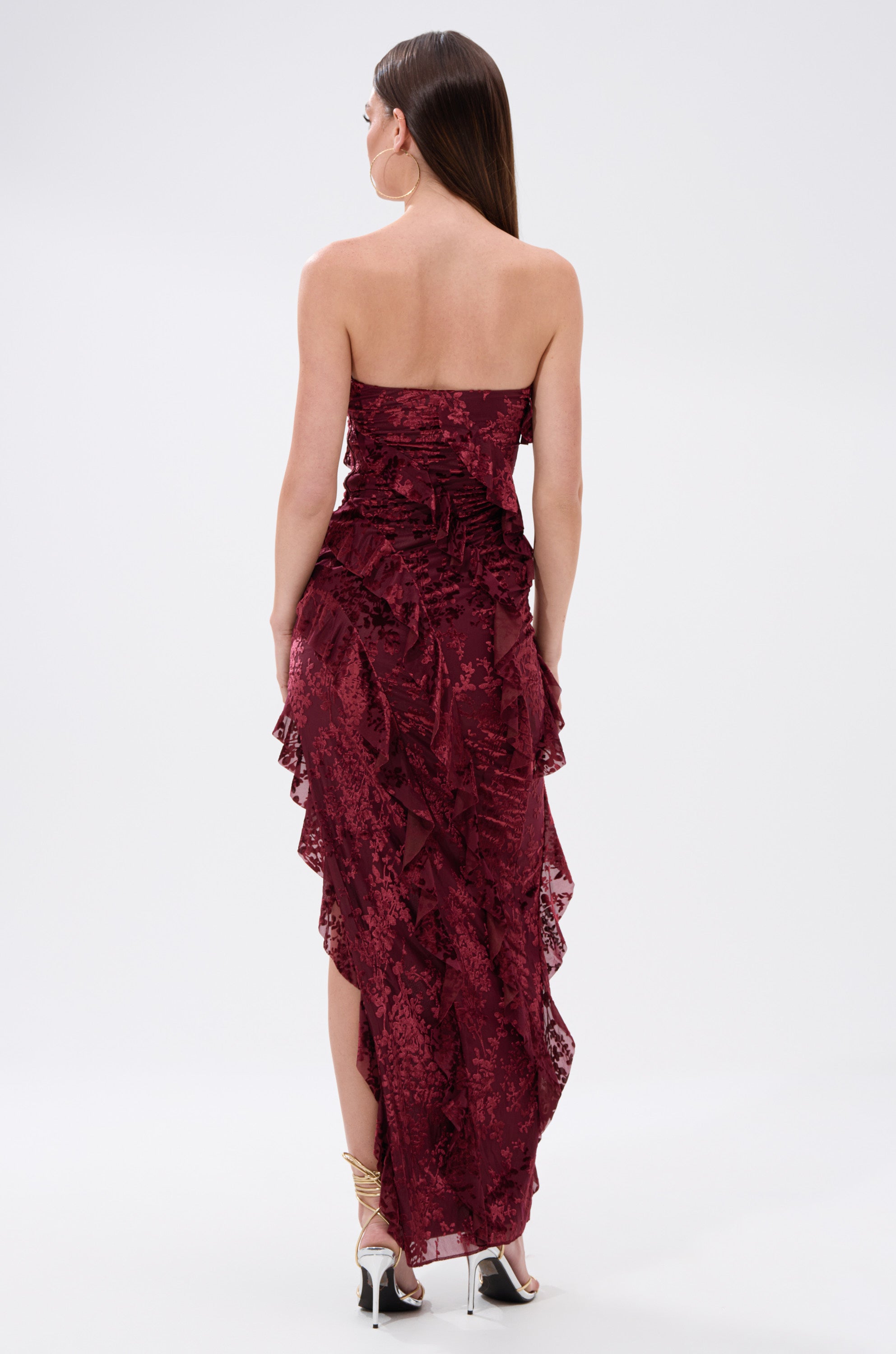 A woman stands with her back to the camera, wearing the WHERE WE GOIN VELVET BURNOUT MIDI DRESS—strapless, dark red velvet with ruffle details and an asymmetrical hem—paired with strappy high-heeled sandals.