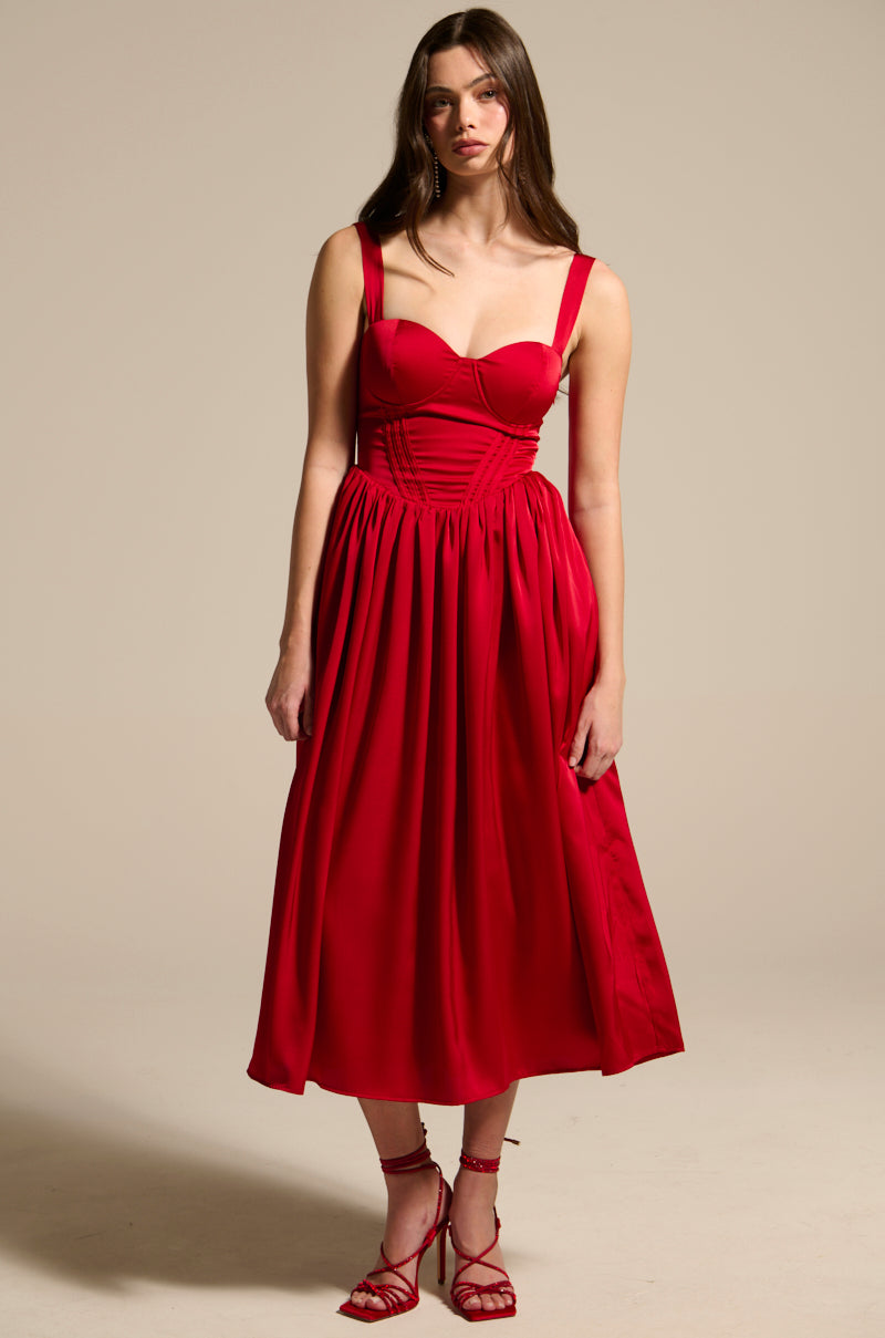 A woman poses against a neutral backdrop in the DEEP INSIDE MIDI DRESS. The red satin dress features a lace-up back, fitted bodice, and full skirt. She completes the look with red strappy heels, while her long hair flows past her shoulders.