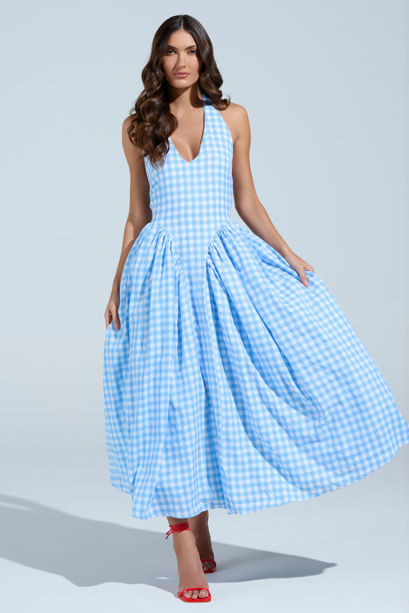 A woman poses in a studio wearing the NEVER GONNA STOP PLAID MAXI DRESS, showcasing its sleeveless design and full skirt. Her long, wavy brown hair flows down her back, and she's paired it with red high heels against a simple light blue backdrop.