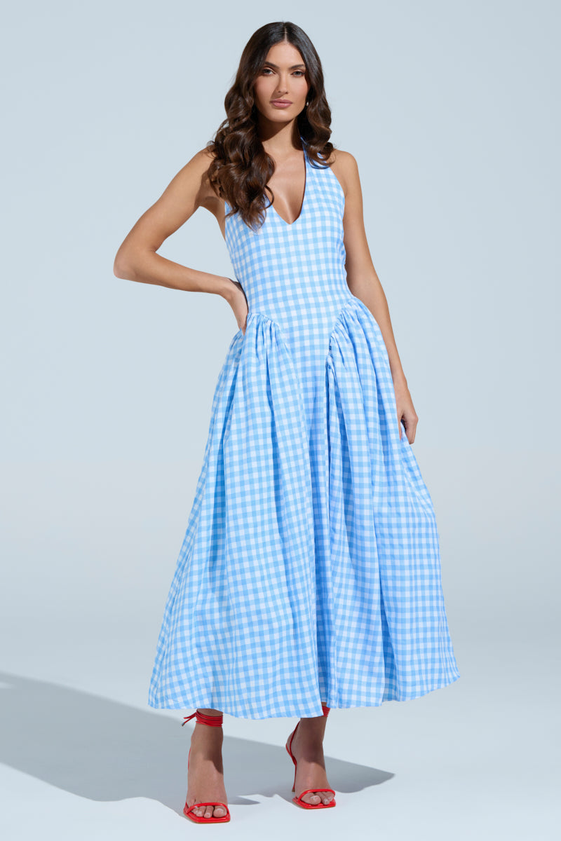A woman stands confidently in the NEVER GONNA STOP PLAID MAXI DRESS featuring a flowing blue and white gingham pattern and deep V-neckline, paired with red high heels. Her long, wavy hair cascades over her shoulders against a simple, light blue background.