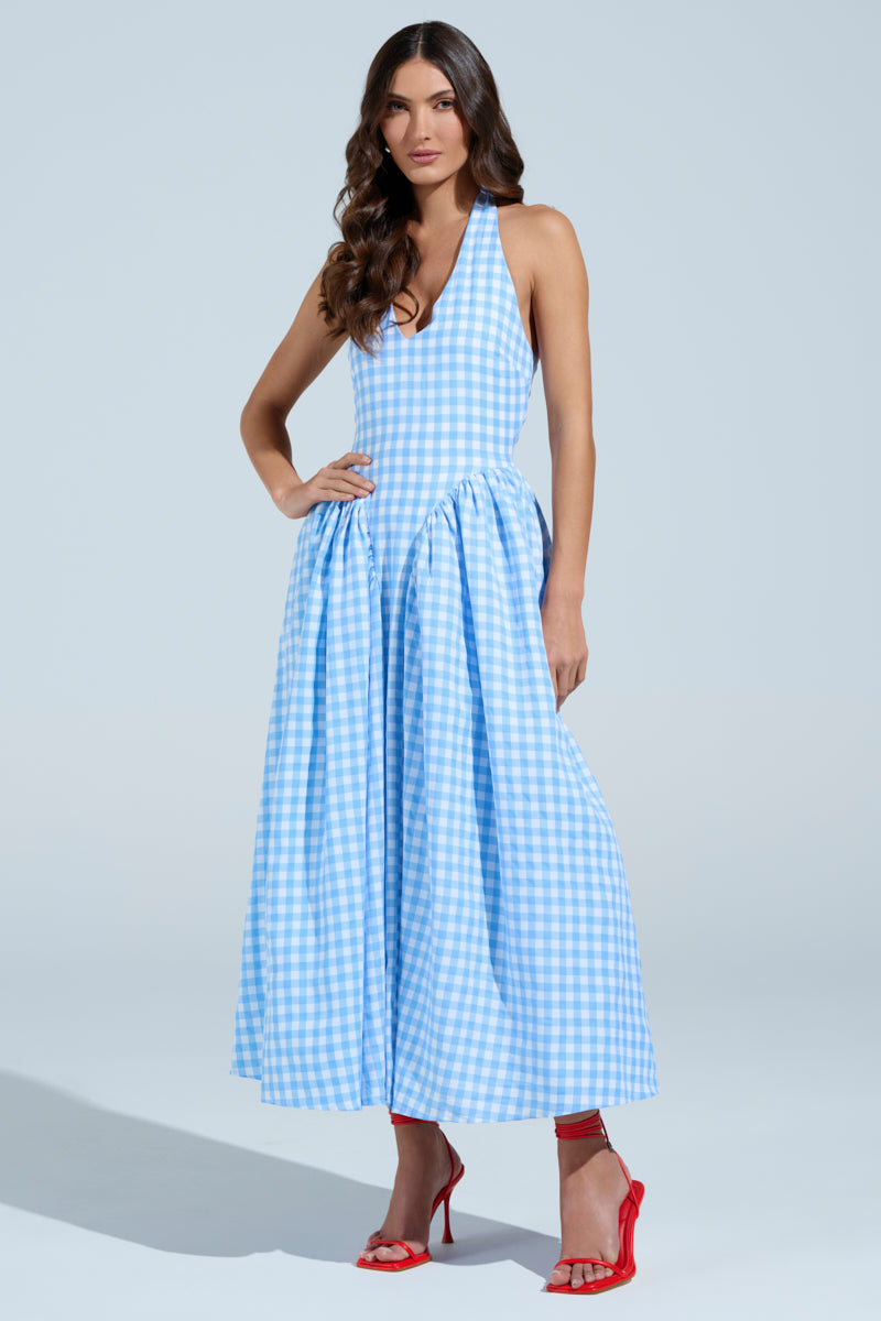 In a light gray backdrop, a woman poses in the NEVER GONNA STOP PLAID MAXI DRESS, featuring blue and white plaid with a sleeveless fitted top and flared skirt. She completes her summer-ready look with red high heels and long, wavy brown hair.