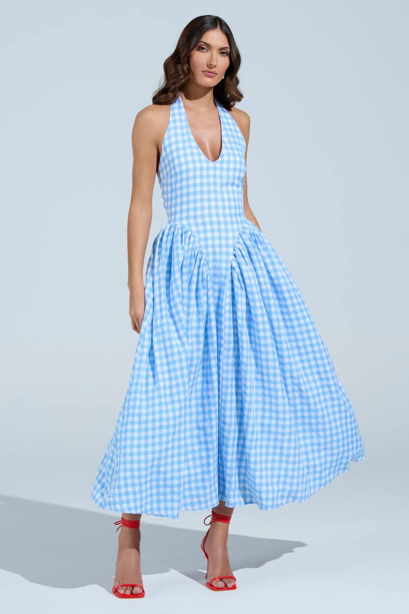 A person poses in the NEVER GONNA STOP PLAID MAXI DRESS featuring blue and white checks with halter straps and a full skirt, elegantly paired with red high-heeled sandals. They stand against a plain light blue background, radiating summer dress vibes.