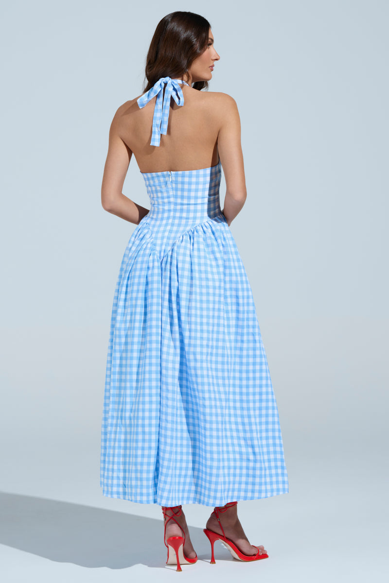 A woman faces away, modeling the NEVER GONNA STOP PLAID MAXI DRESS—a blue and white gingham summer piece with a fitted bodice and full skirt. Halter straps accent her long hair, paired with her red high-heeled sandals against a plain light gray background.