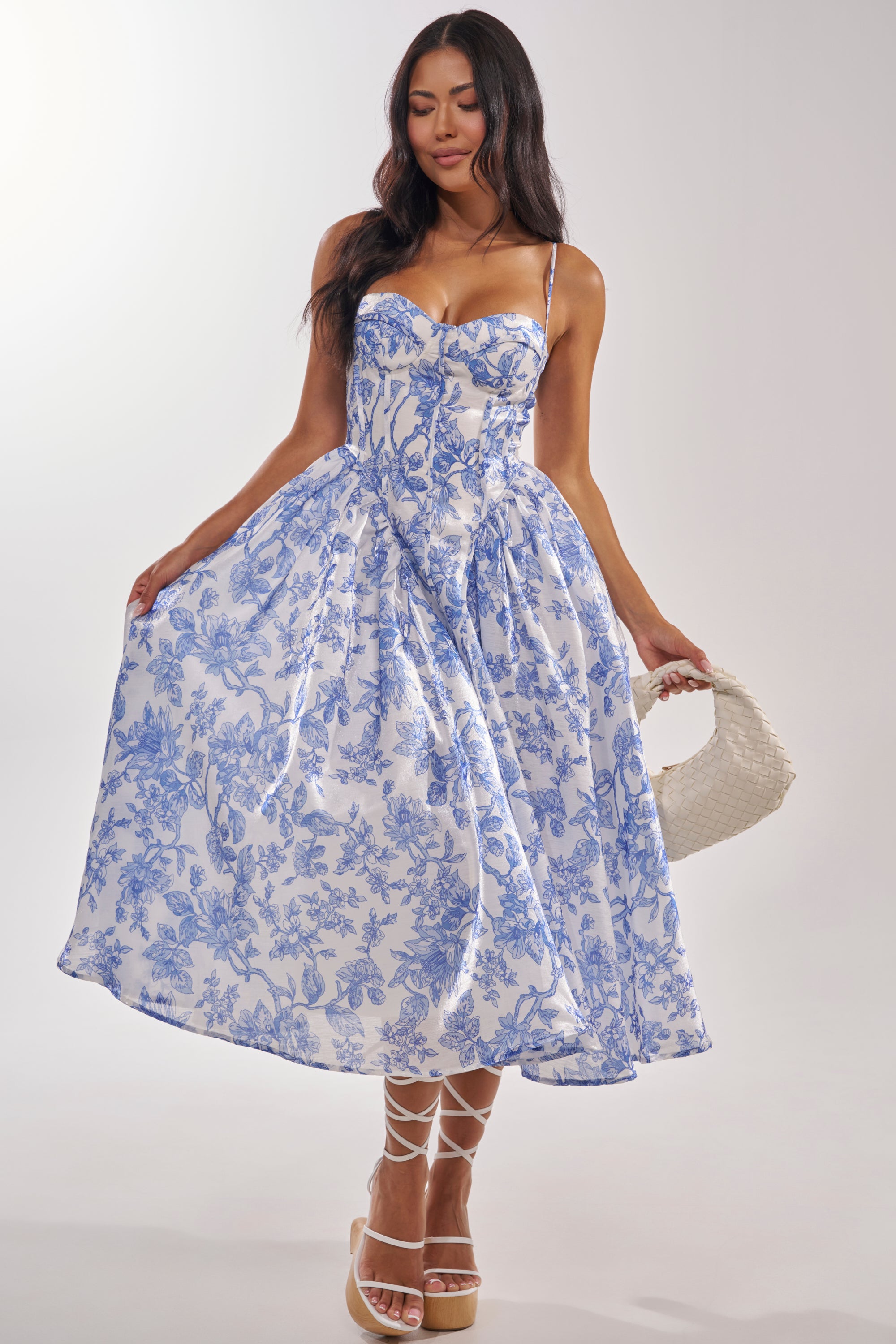 A woman models the FEELING FLORAL MAXI DRESS, a white gown with blue floral patterns, strappy sandals, and a textured handbag. The dress has a fitted bodice, thin straps, and a full skirt—ideal for formal occasions. She stands against a plain background. 4