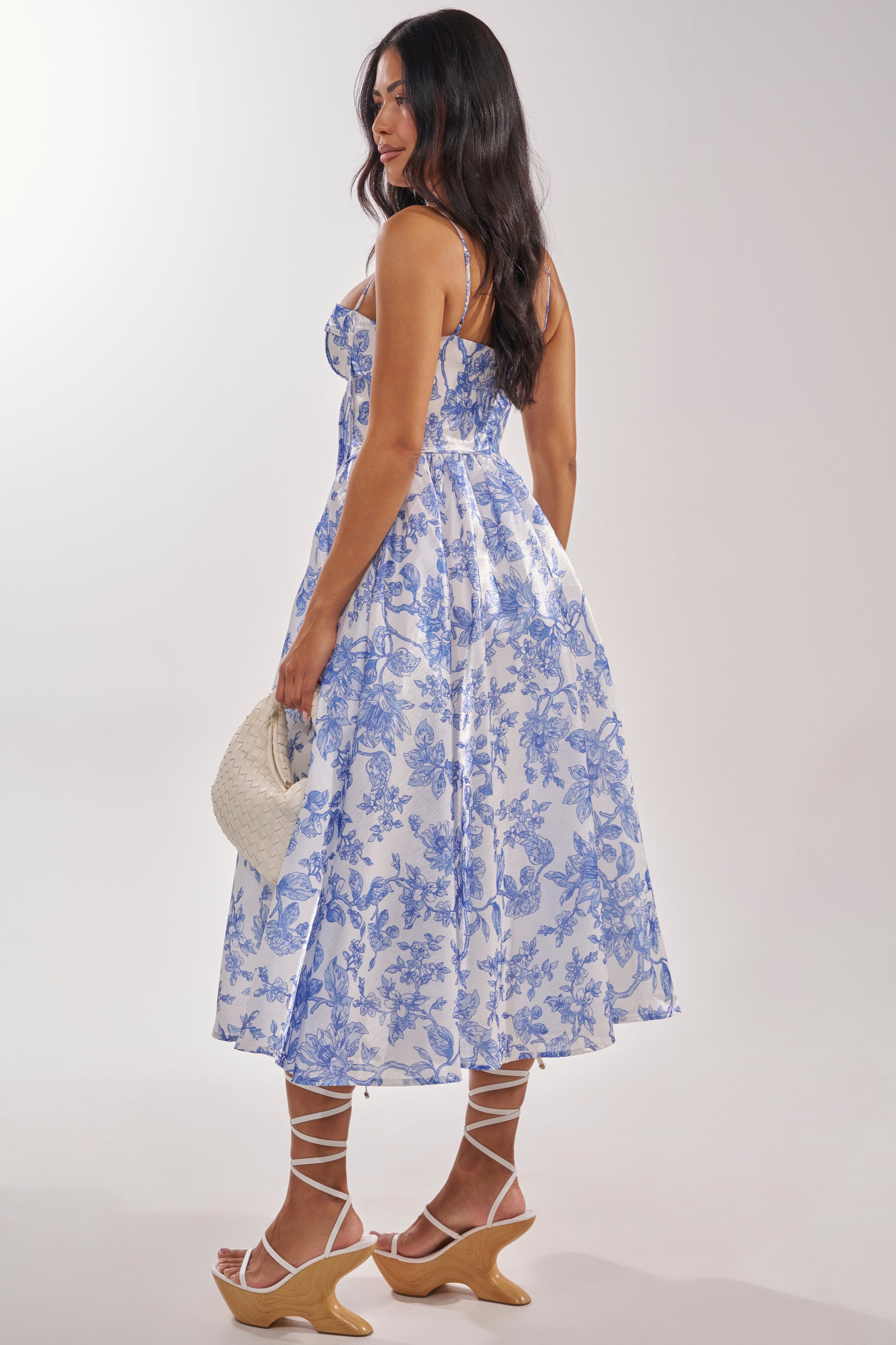 A woman in profile wears the FEELING FLORAL MAXI DRESS with spaghetti straps, lace-up sandals, and a woven white handbag. With long dark hair and a soft smile, she looks perfectly styled for any formal occasion. 5
