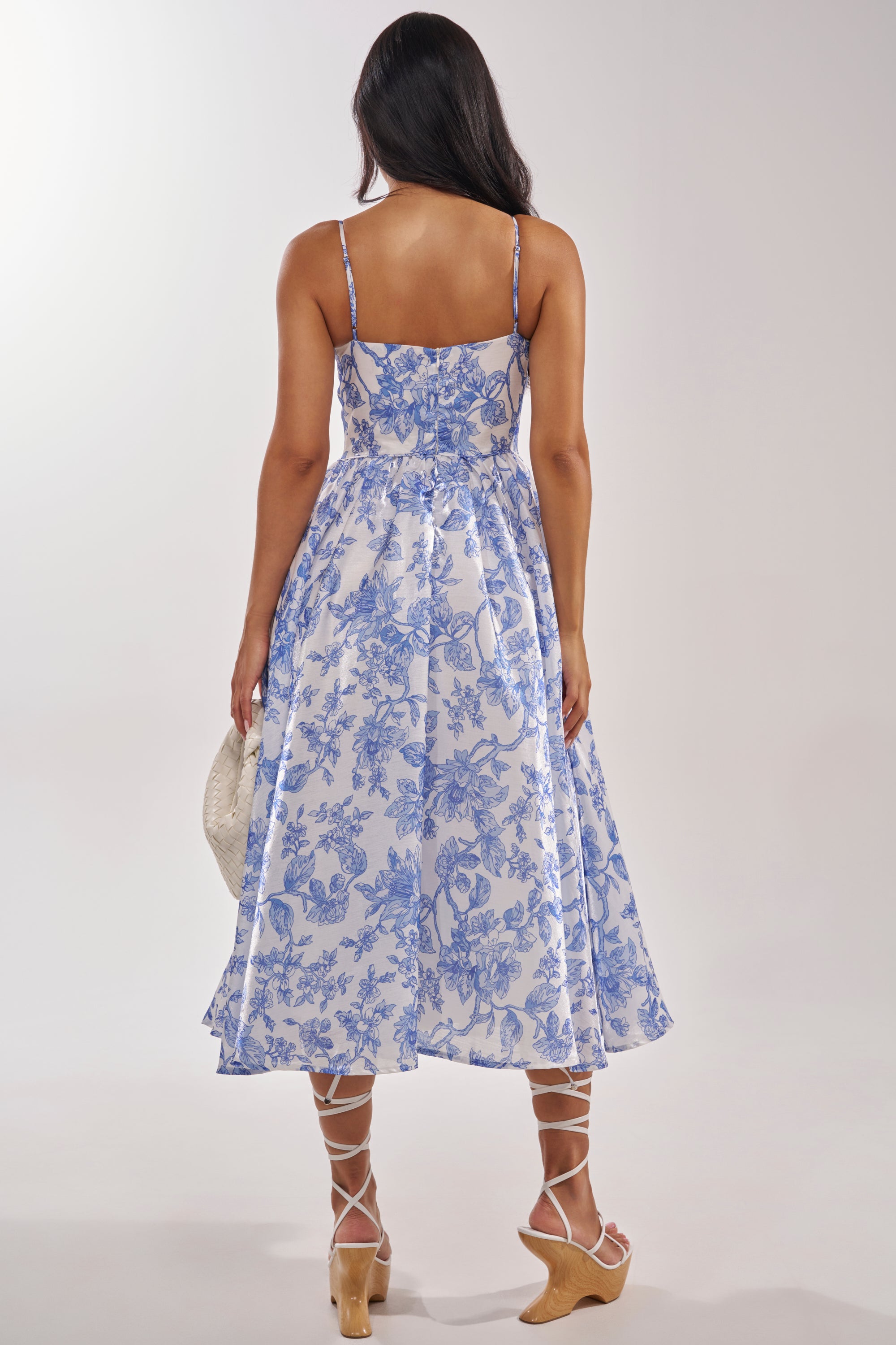 A woman with long dark hair, seen from behind, wears the FEELING FLORAL MAXI DRESS with strappy sandals and holds a light woven hat. The plain background highlights the dress's blue floral print. 6