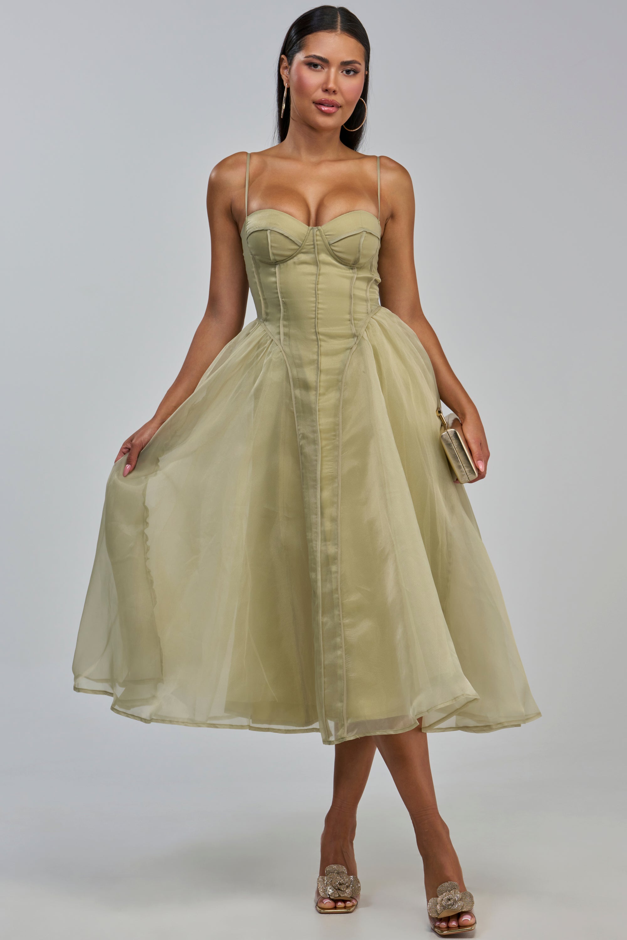 Wearing the COLETTE MAXI DRESS IN OLIVE—a light olive green, corset-style, tea-length tulle dress with thin straps—she holds a small clutch and wears open-toe heels, posing elegantly against a plain background; it’s the perfect choice for a wedding guest dress.