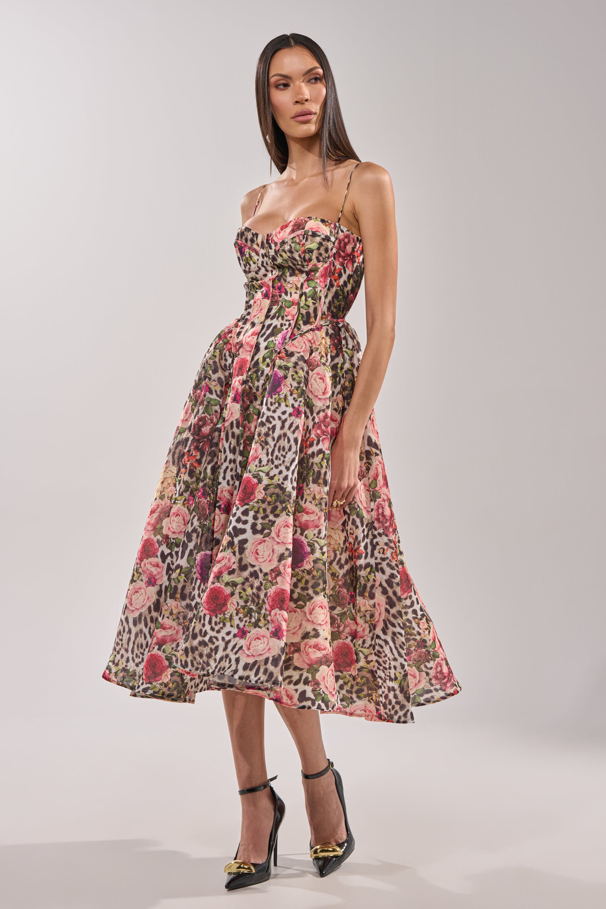 A woman models the COLETTE MAXI DRESS, featuring a sleeveless leopard print design with thin straps and a fitted corset bodice. She pairs it with black heels and stands against a plain, light gray background. 0