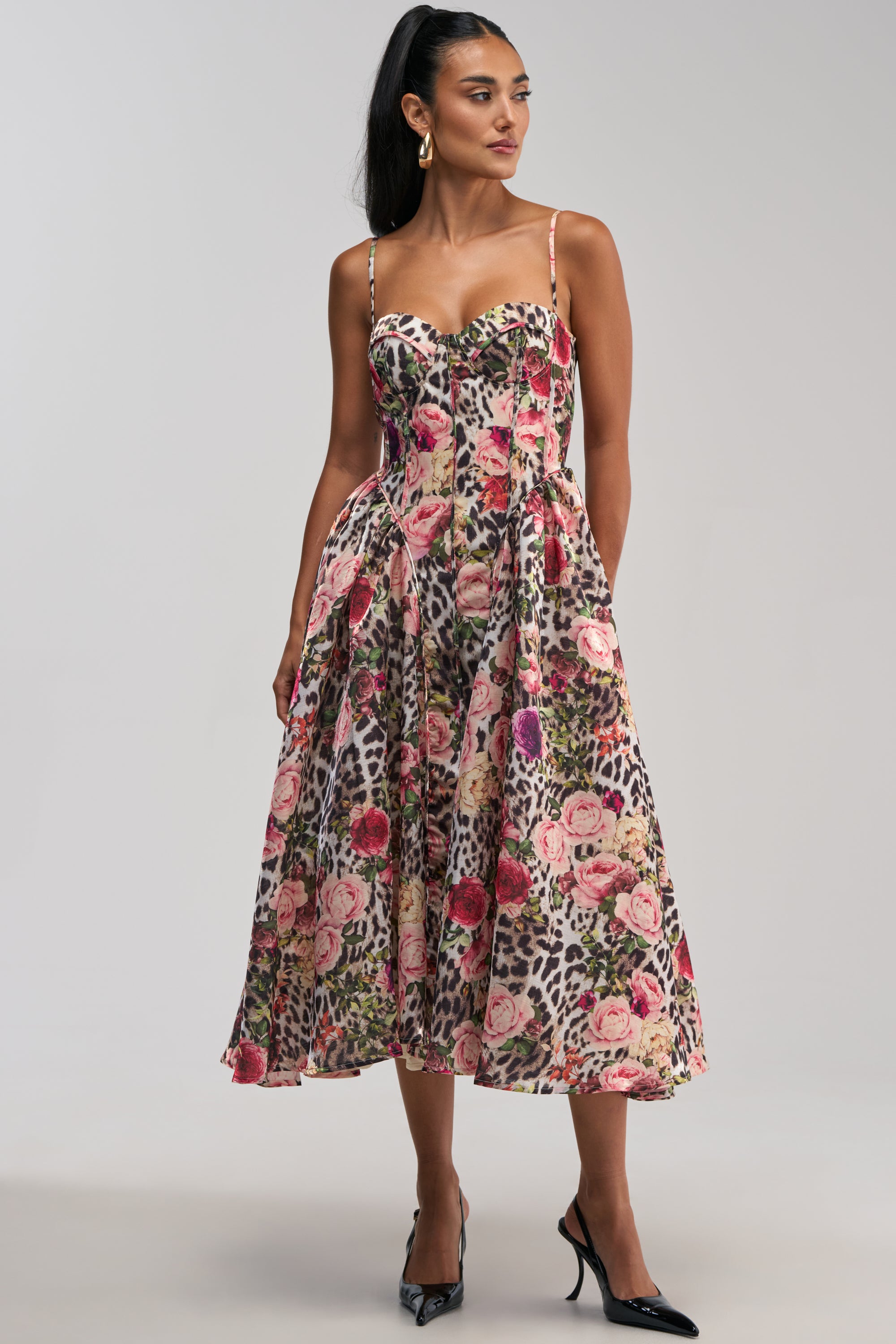 A woman wearing the COLETTE MAXI DRESS, a sleeveless, mid-length dress with a pink floral and leopard print pattern, featuring a flattering sweetheart neckline and fitted bodice with flared skirt. She has long dark hair in a ponytail and accessorizes with black heels and gold hoop earrings.